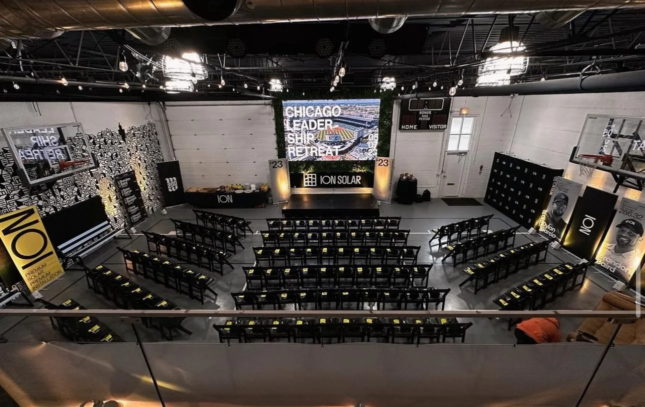 An indoor conference or event space arranged with rows of black chairs with yellow napkins, facing a stage with a large screen, banners, and signs displaying 'ION Solar' and related phrases.
