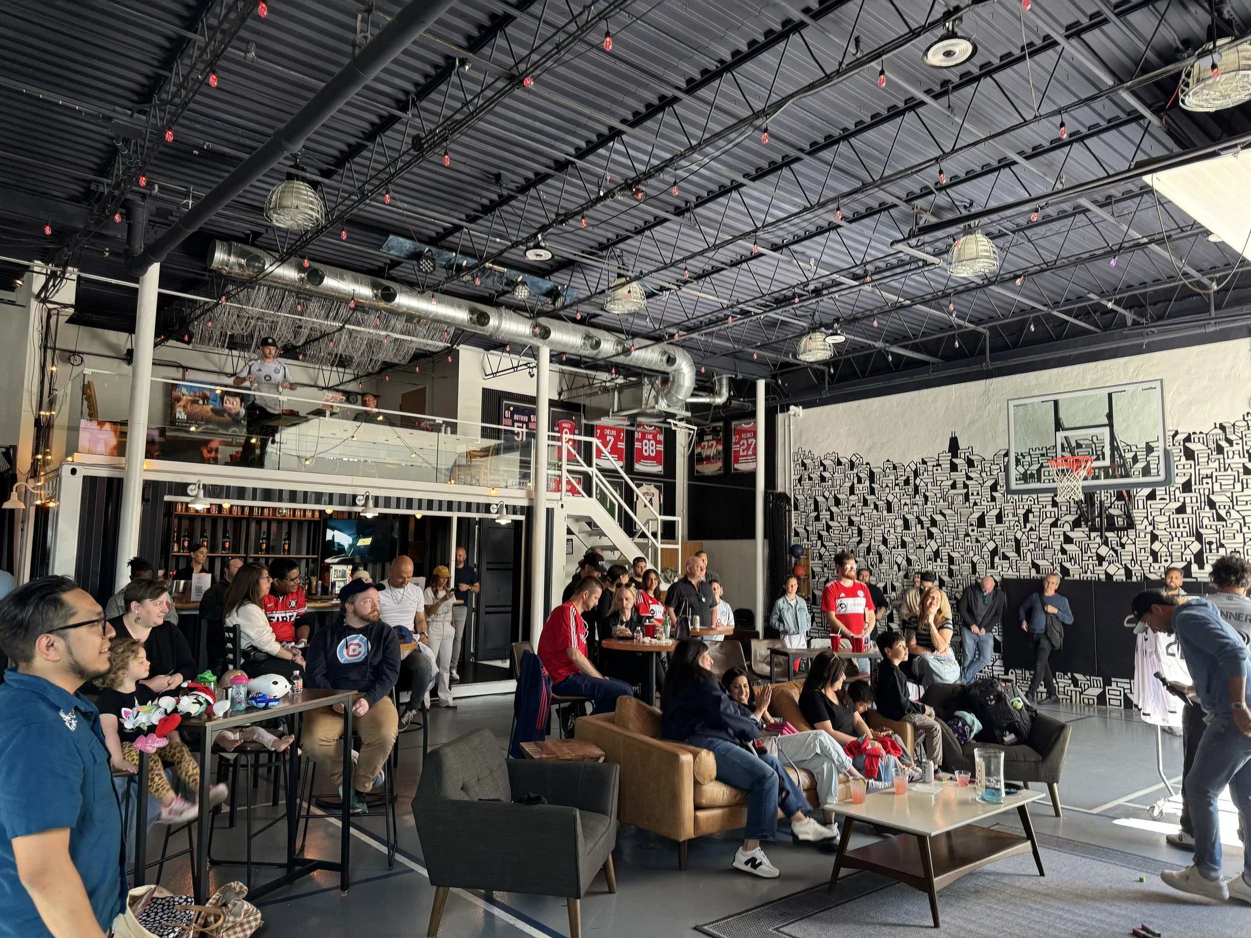 Indoor scene at a sports bar or restaurant filled with people watching a game or event on TV screens. The ceiling has exposed industrial ductwork and lighting fixtures. Patrons are seated on various chairs and sofas, some holding drinks and snacks, w