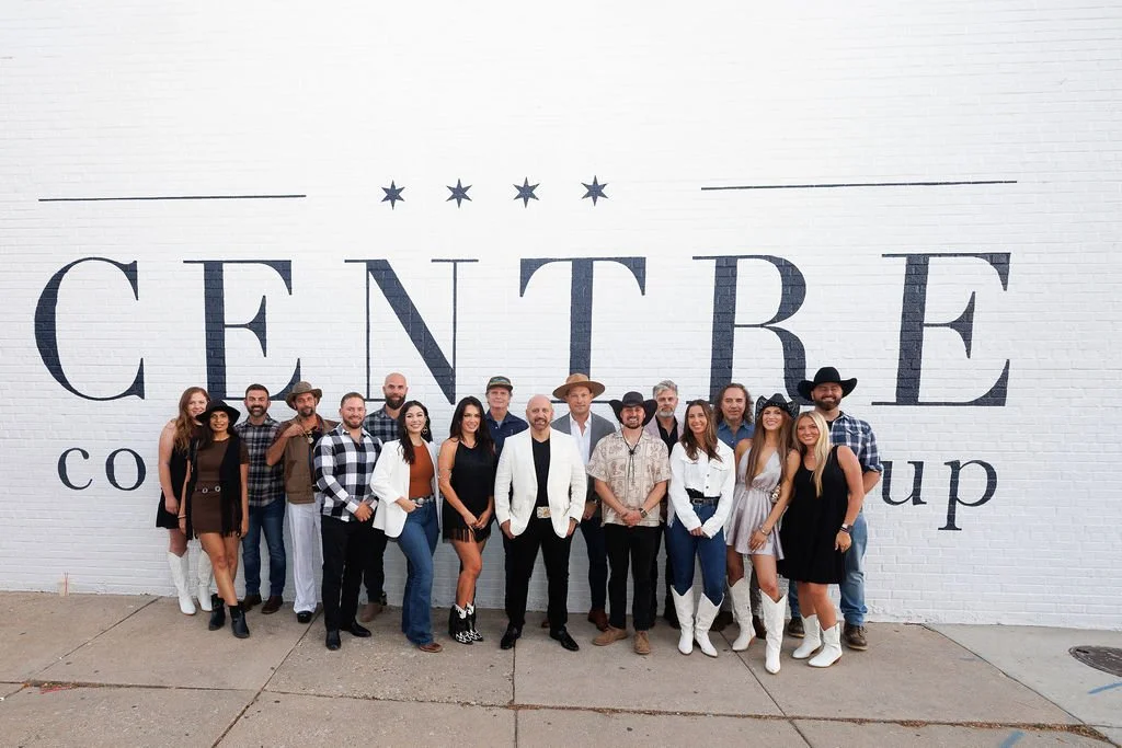 Group of people standing in front of a white brick wall with large text reading 'CENTRE'. They are dressed in a mix of casual and cowboy-themed attire, with some wearing cowboy hats and boots.