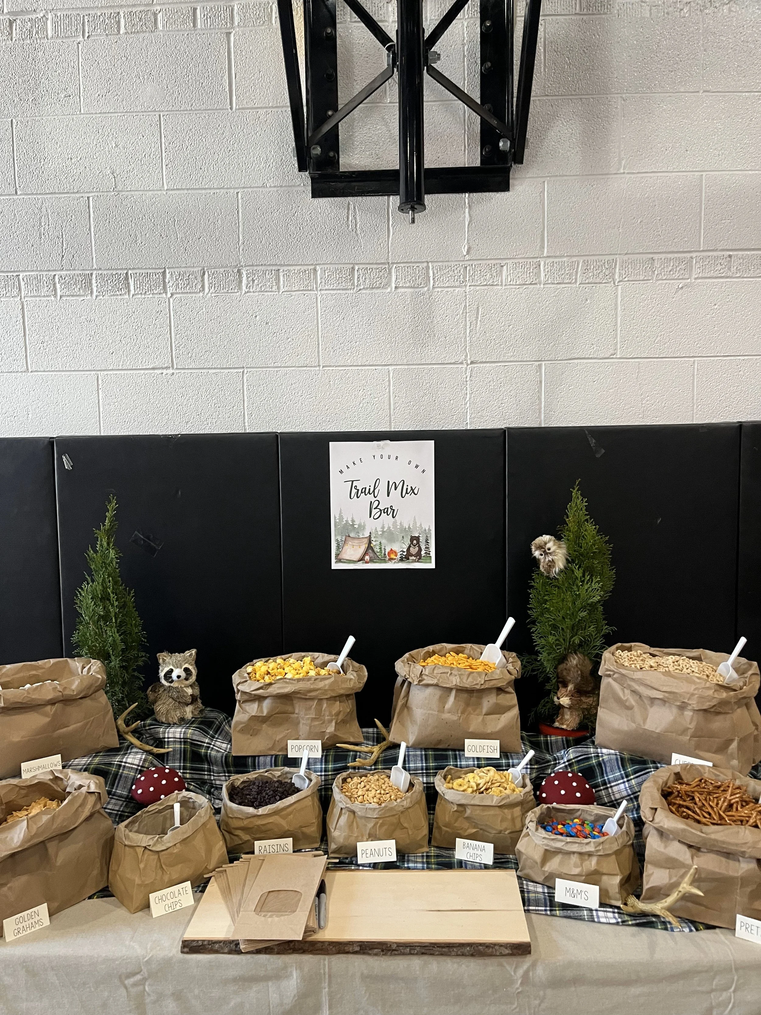 A display of various trail mix ingredients in brown paper bags with labels, including marshmallows, popcorn, raisins, peanuts, goldfish, chocolate chips, M&M's, pretzels, and banana chips, with small decorative trees and a sign reading 'Make Your Own
