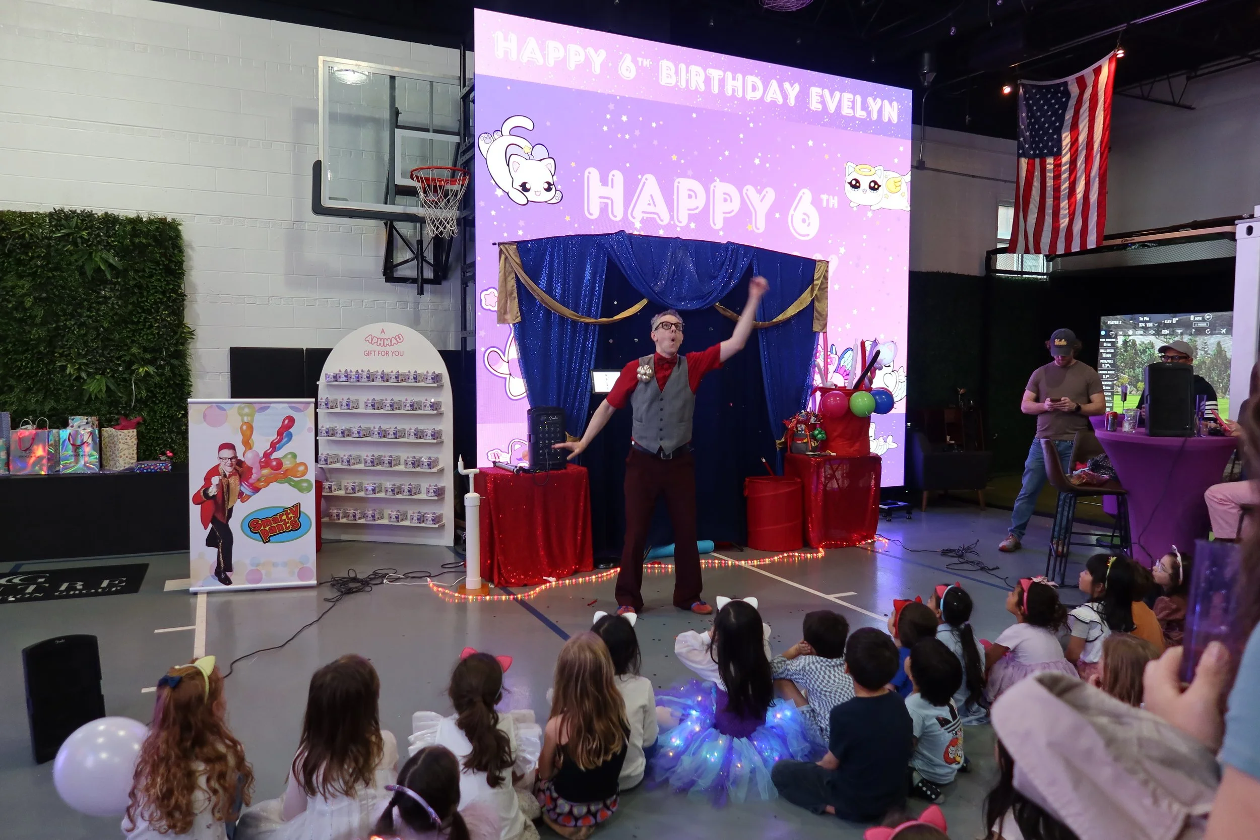 Children sitting on the floor watching a magician performing on stage at a birthday party celebration. The stage has a large pink backdrop with animated characters and the text 'Happy 6th Birthday Evelyn.' There is a man in a red shirt and gray vest 