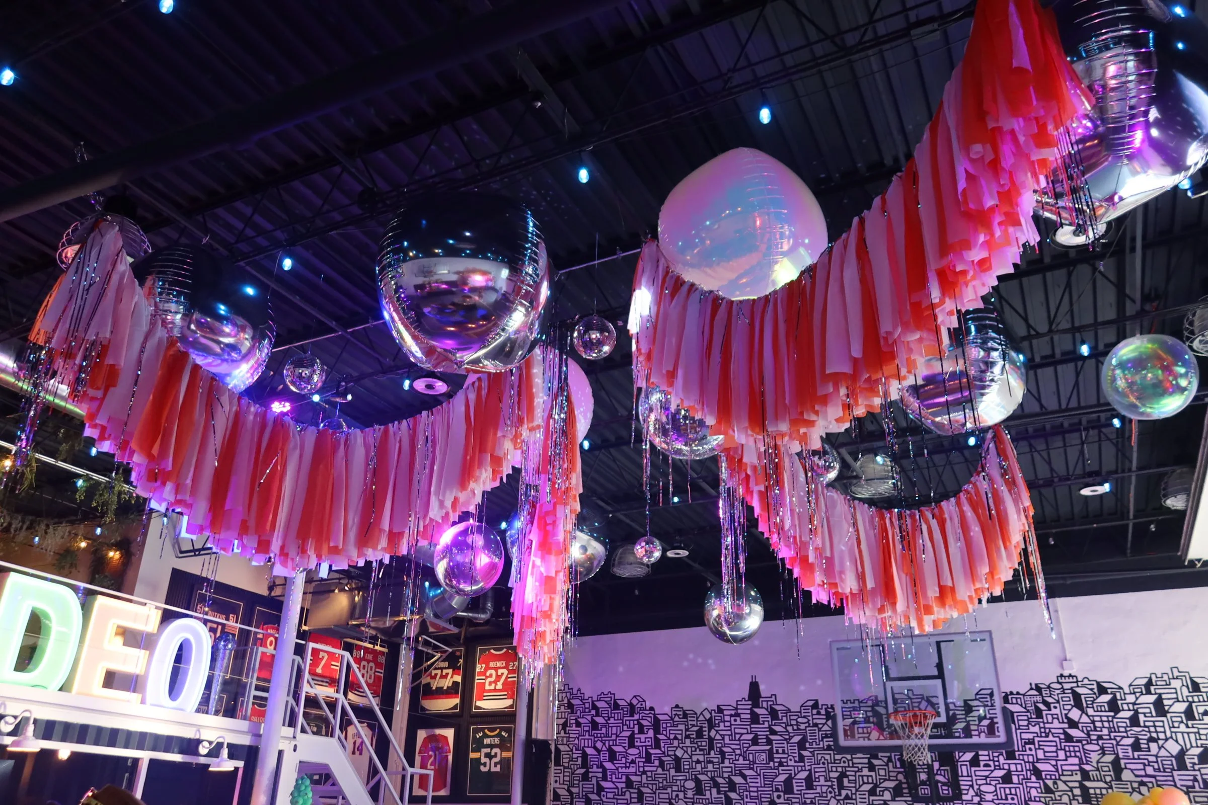 Colorful hanging decorations with pink streamers and reflective silver orbs hanging from a ceiling in a lively indoor setting.