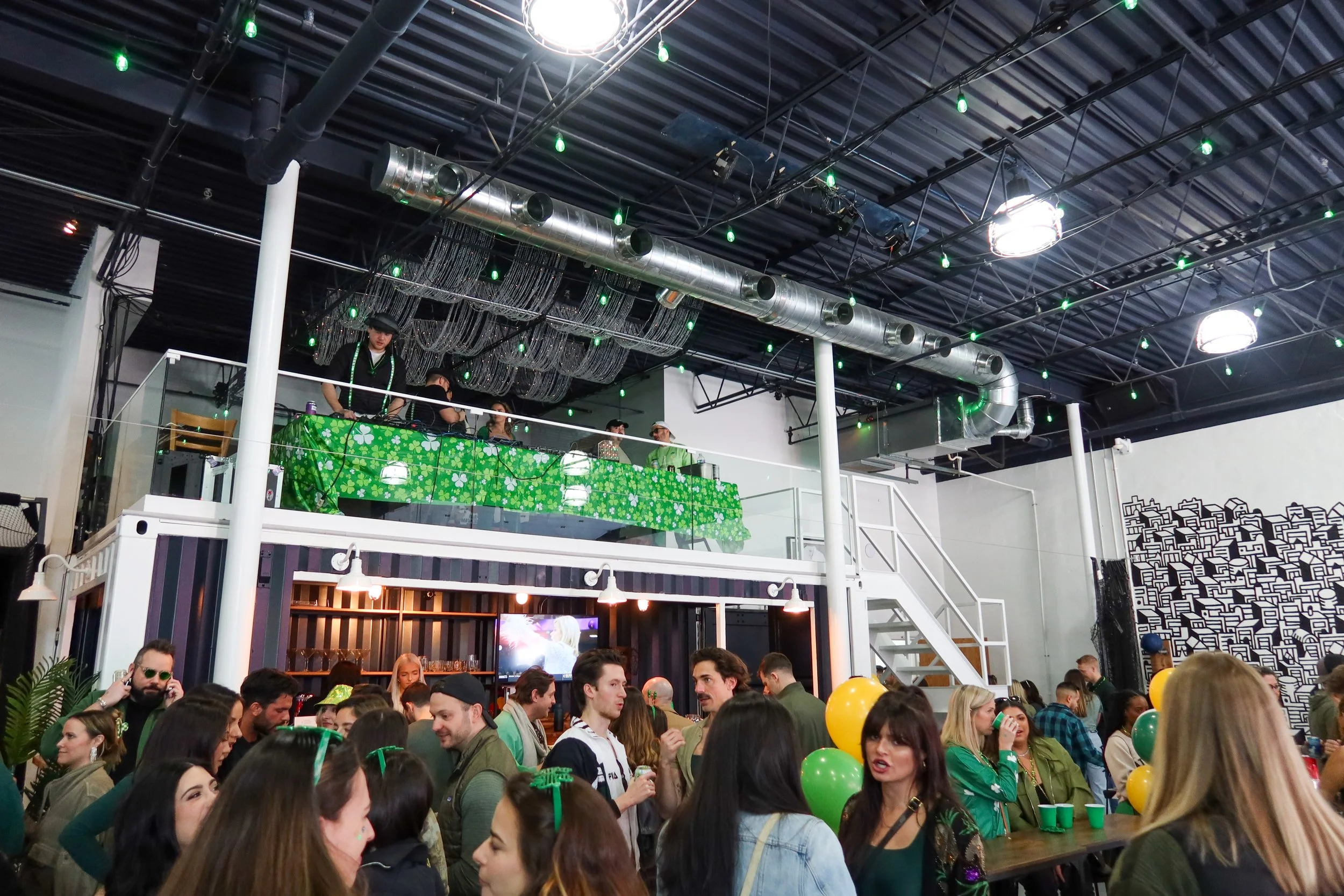 Crowd of people celebrating with green balloons and St. Patrick's Day decorations inside a festive indoor venue with green and yellow decor.