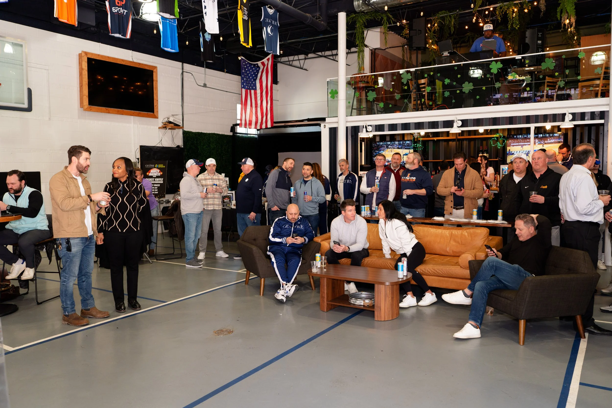 People gathered inside a sports bar decorated for St. Patrick's Day with shamrock banners, American flags, and sports jerseys hanging from the ceiling. Some individuals are standing and chatting, while others are seated and looking at their phones.