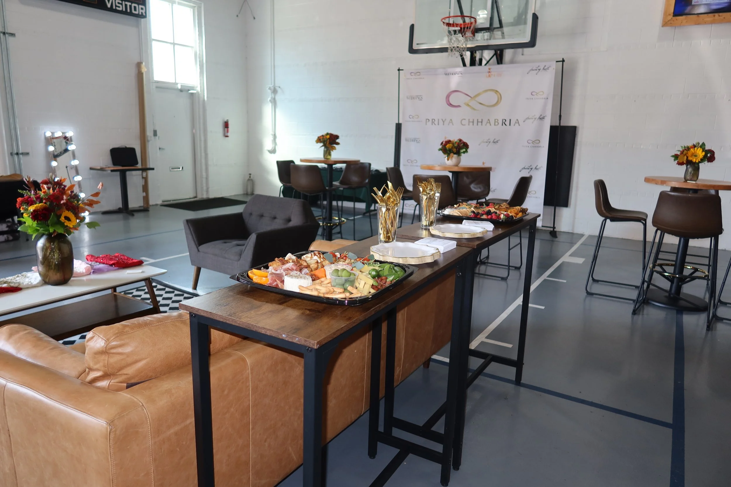 Indoor event setup in a gymnasium with tables of food, flower arrangements, and a backdrop with a logo for Priya Chhabria.