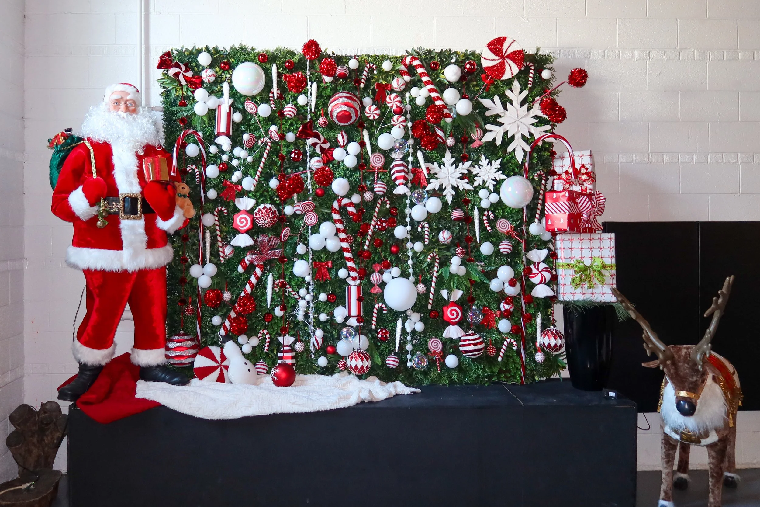 Santa Claus dressed in red and white, standing next to a decorated Christmas tree with red, white, and silver ornaments, candy canes, and large snowflakes, with wrapped gifts at the base, a reindeer figure to the right, and a white wall in the backgr