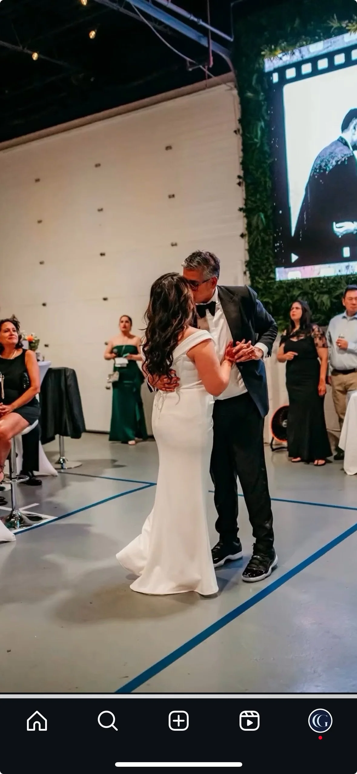 Couple dancing at a wedding reception, with guests watching in the background and a large screen displaying an image of the couple.