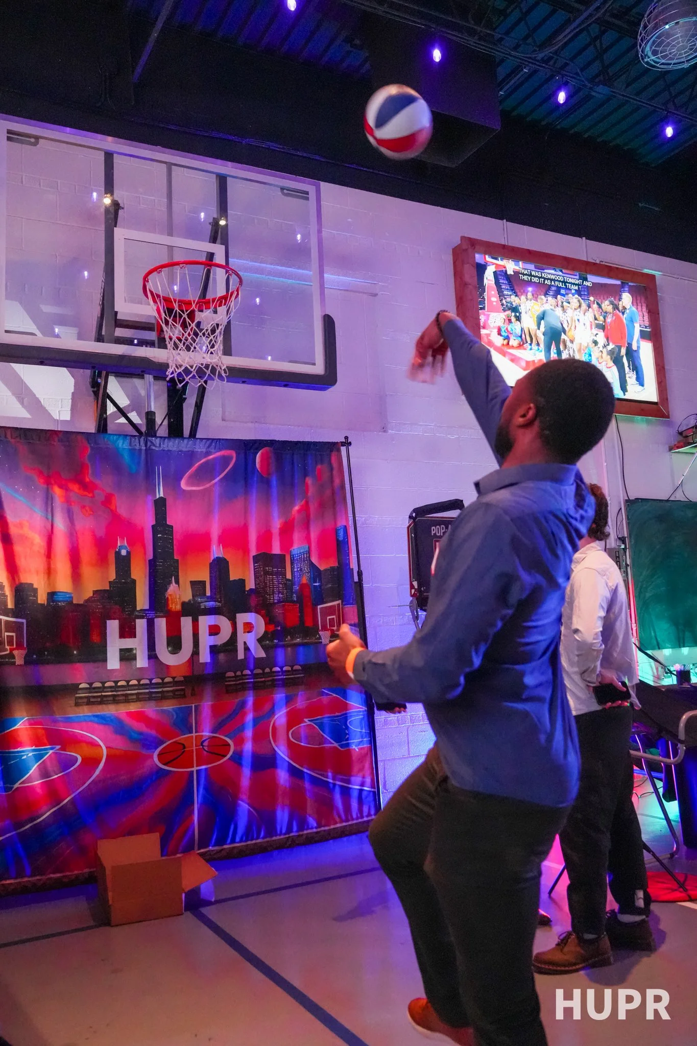 People playing arcade basketball game in an arcade with a basketball hoop, a screen showing a retro cityscape, and a TV displaying a sports broadcast.
