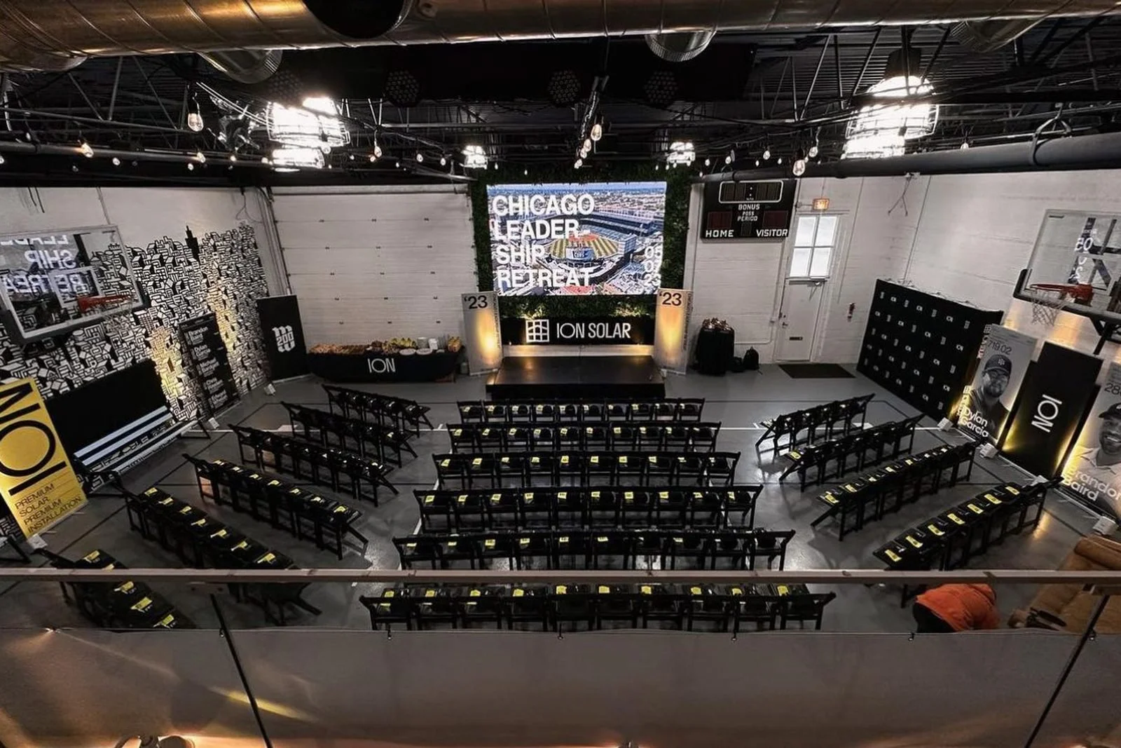 Indoor event space with chairs arranged facing a stage with a large screen displaying text about Chicago leadership and retreat. The room has advertisements for Ion Solar, a scoreboard, and a basketball hoop on the left wall, with dim lighting and a modern industrial decor.