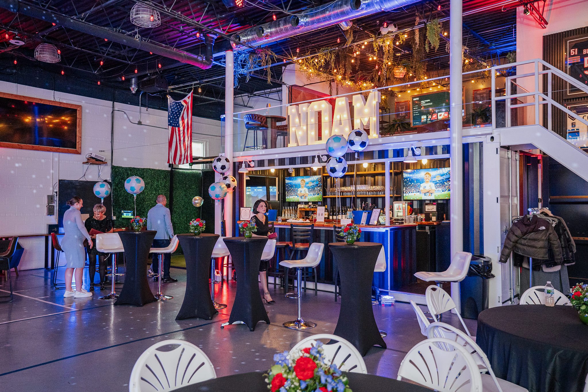 Indoor sports bar decorated for a celebration with hanging soccer ball balloons, floral centerpieces on tables, a large illuminated sign spelling 'FLAM' and multiple flat screen TVs showing a soccer game.