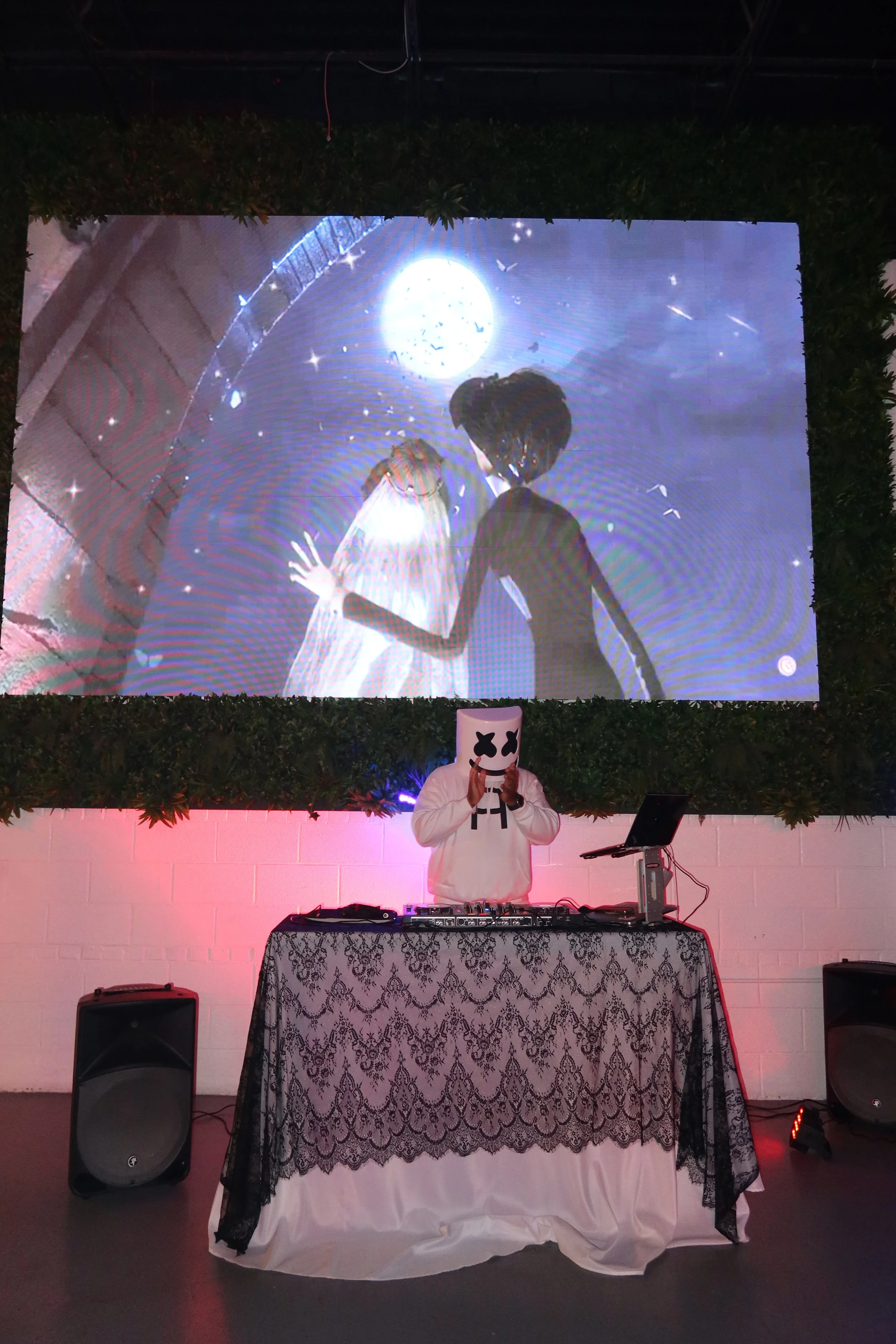 DJ wearing a mask with Xs over the eyes and a striped hoodie, standing behind a DJ booth with turntables, in front of a pink-lit wall, with a large screen displaying an animated scene of a girl looking at the moon and a starry sky.
