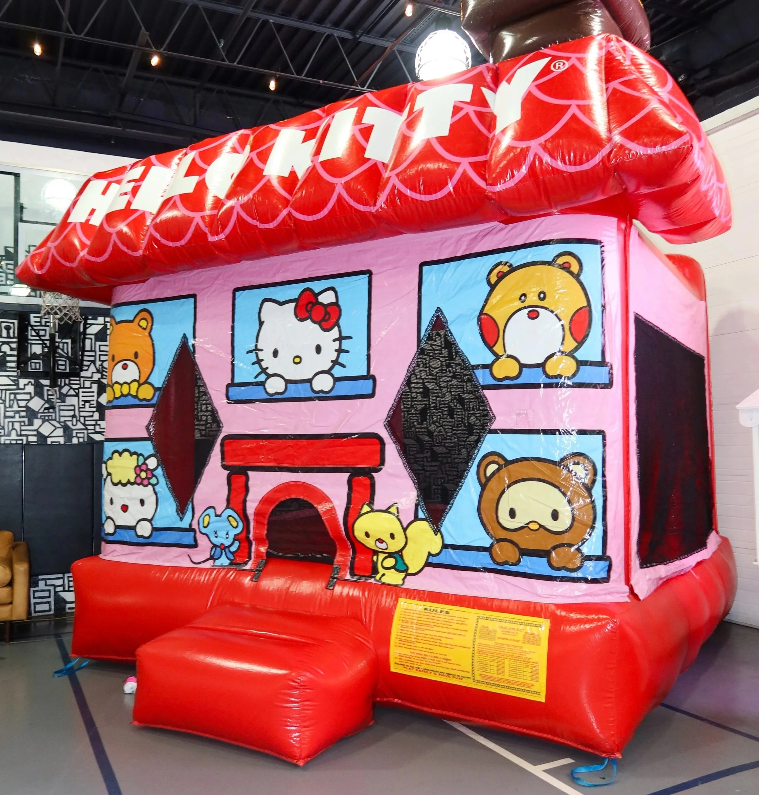 A large pink and red inflatable bounce house with cartoon animal characters and a small red slide, located in an indoor gymnasium.