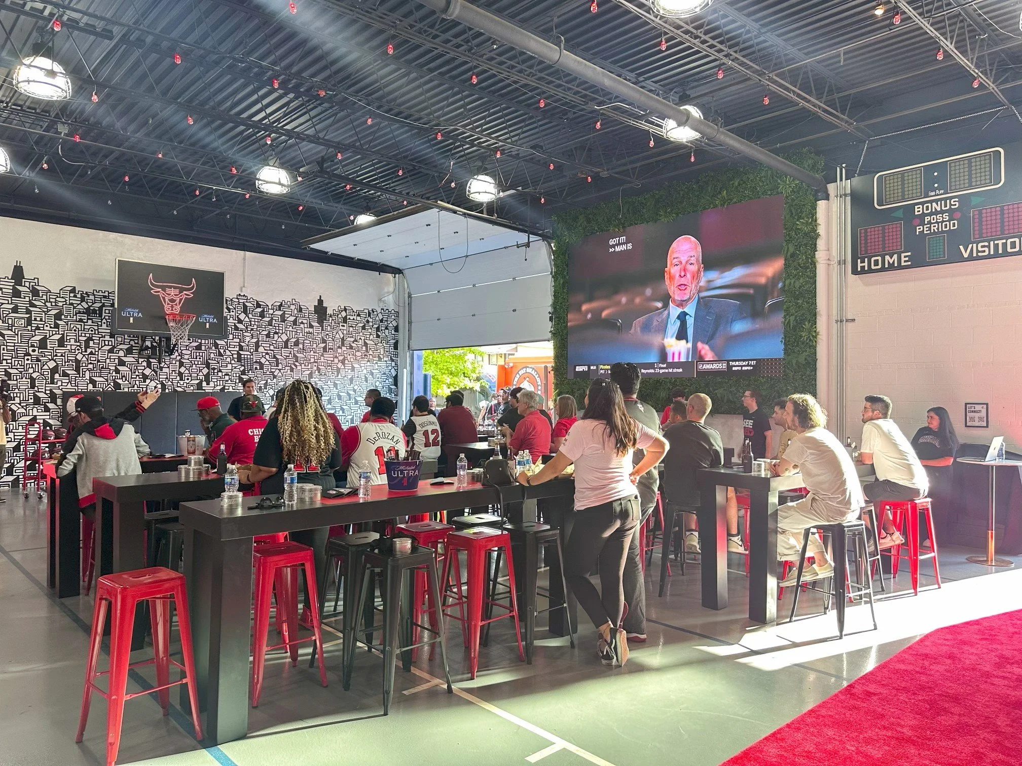 View of a restaurant or sports bar with a crowd of people, a large TV screen showing a man speaking, neon signs, and Chicago Bulls decor, including a basketball hoop and team logos.