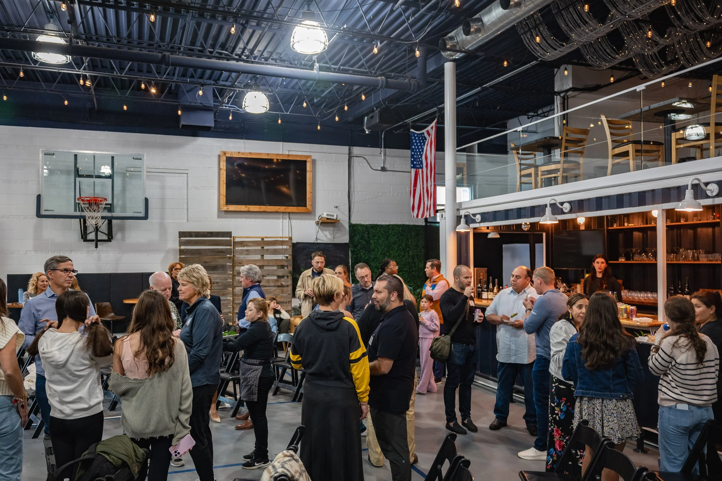 People gathering at a social event or party inside a modern industrial-style venue with high ceilings, exposed pipes, a large flat-screen TV, an American flag, and a bar area.