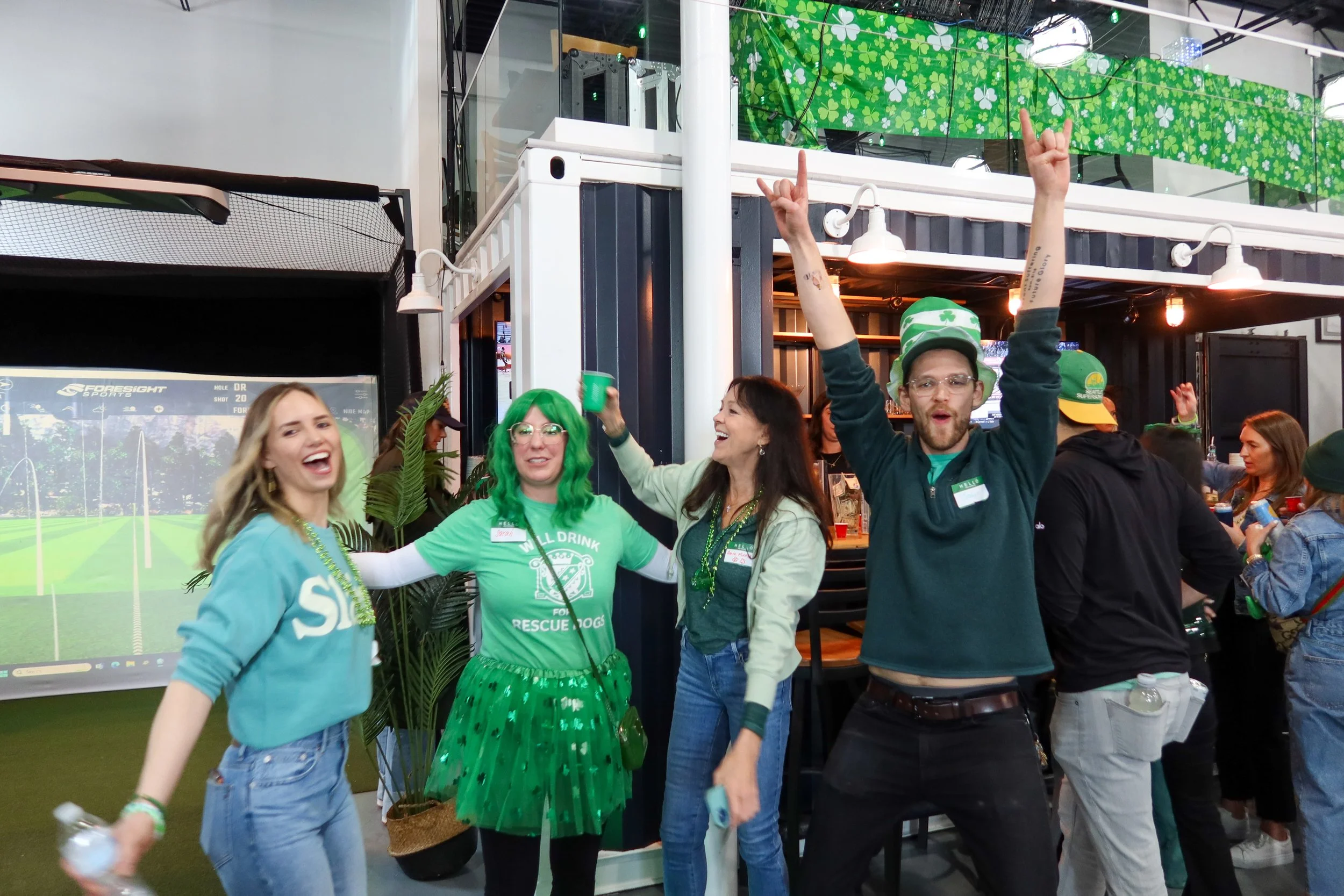 Group of people celebrating St. Patrick's Day with green accessories and clothing, in a lively indoor setting decorated with shamrocks and Irish themes.