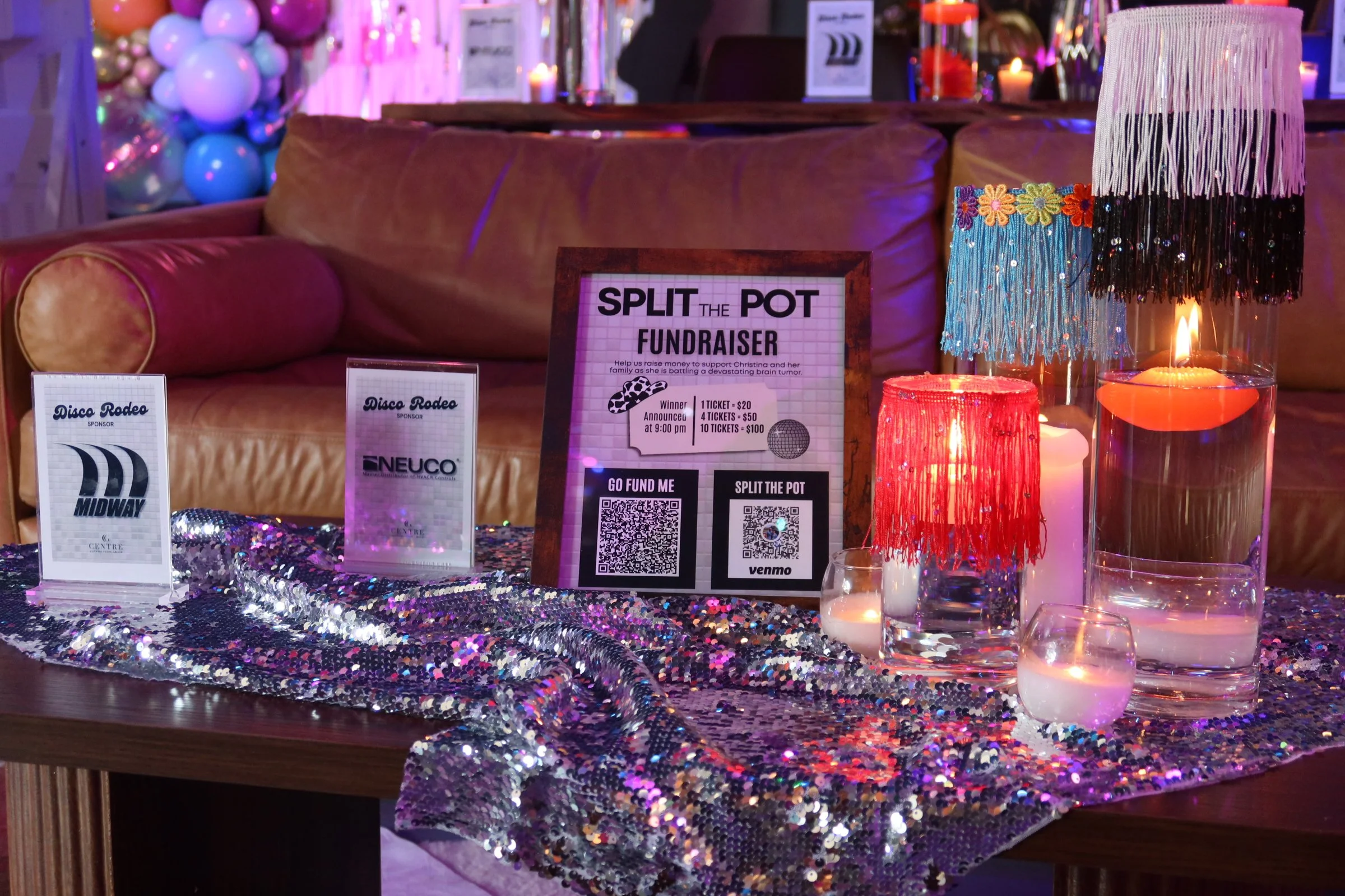 A table decorated with a shiny, sequined tablecloth, featuring event signs for a charity fundraiser and sponsor logos, with colorful lamps and candles, and balloons in the background.