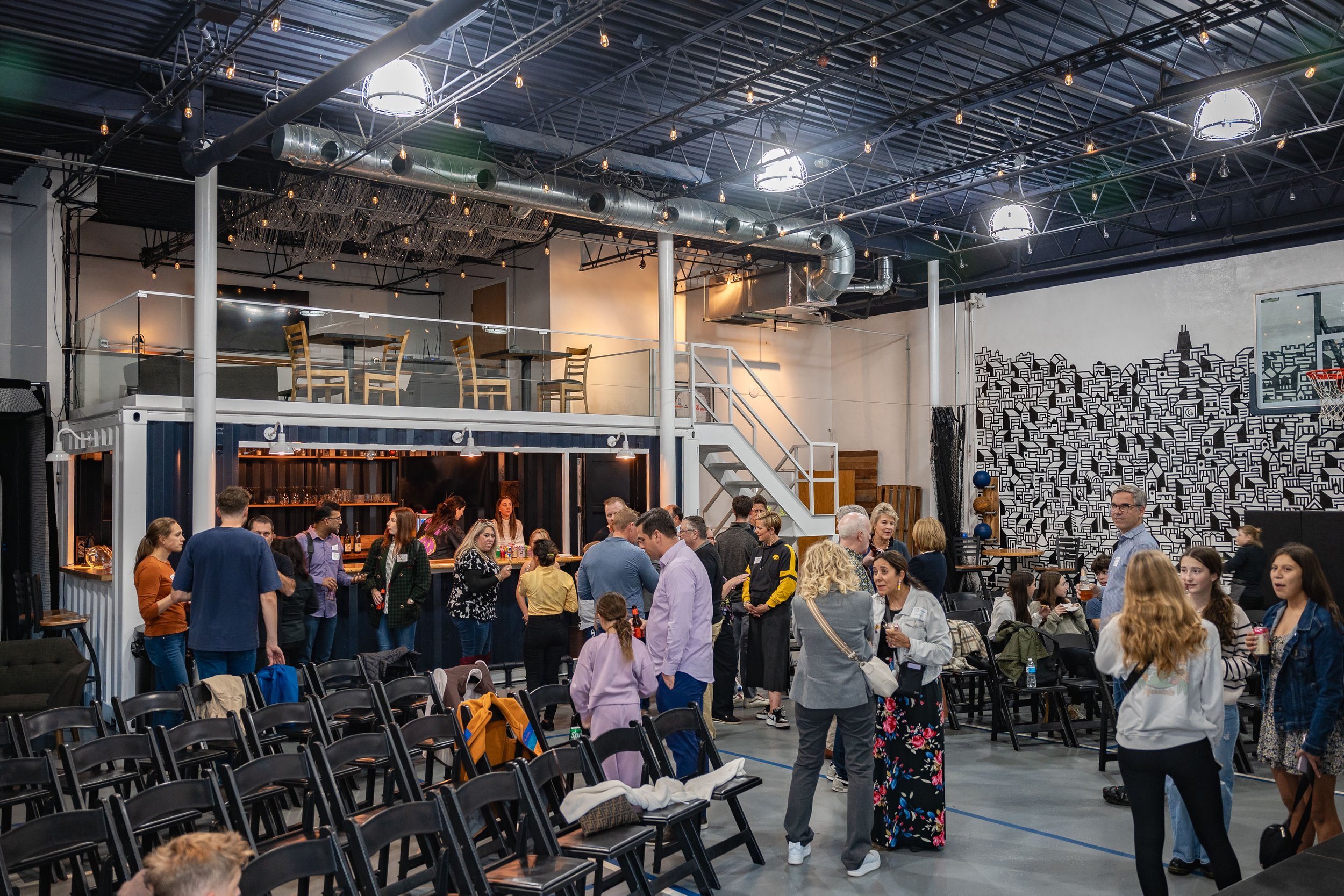 A large indoor gathering space with many people socializing and seated. The space has an industrial style with exposed pipes and strings of lights hanging from the ceiling. There are black chairs, a bar area with glasses, and a wall mural of a citysc