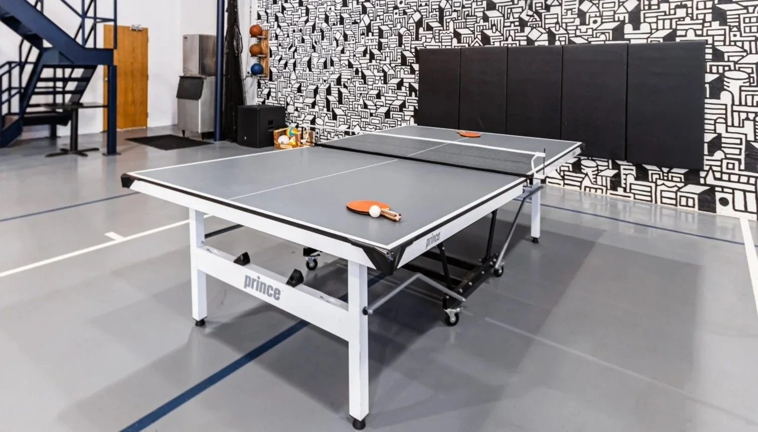 Indoor table tennis setup with two paddles and a ball on a gray table, against a black and white patterned wall, in a recreational space.
