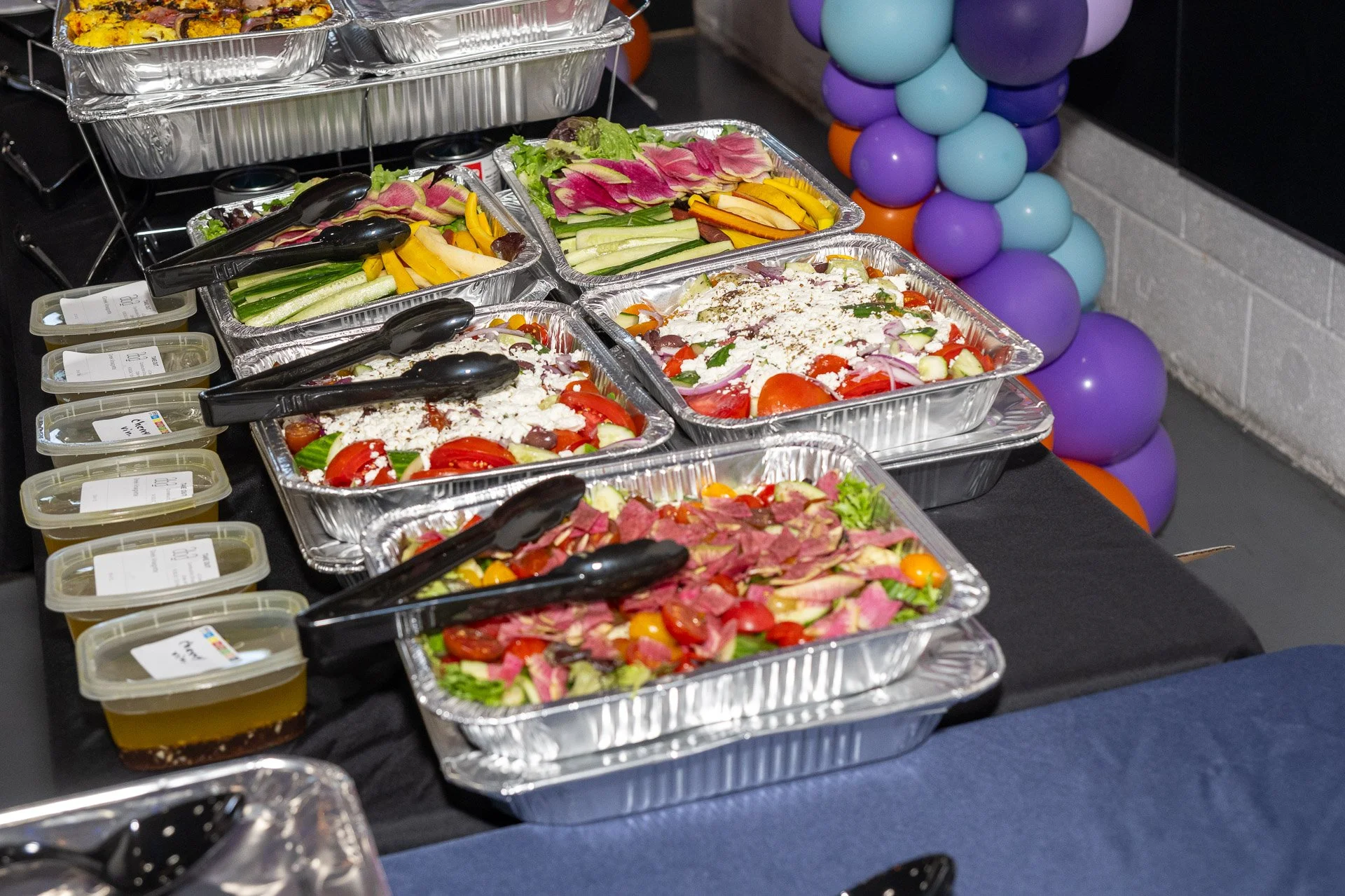 Trays of salad with chopped vegetables, cheese, and toppings on a table decorated with balloons.