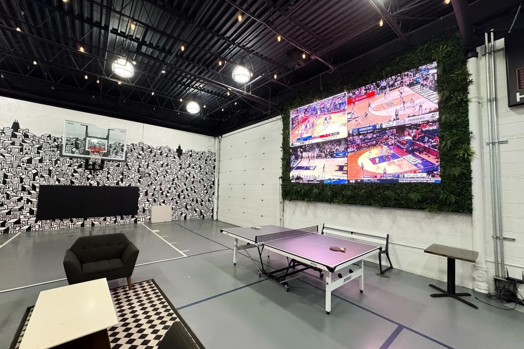 Indoor sports area with black and white geometric wall art, a basketball hoop, a ping pong table, and multiple large screens displaying basketball games, illuminated by ceiling lights.
