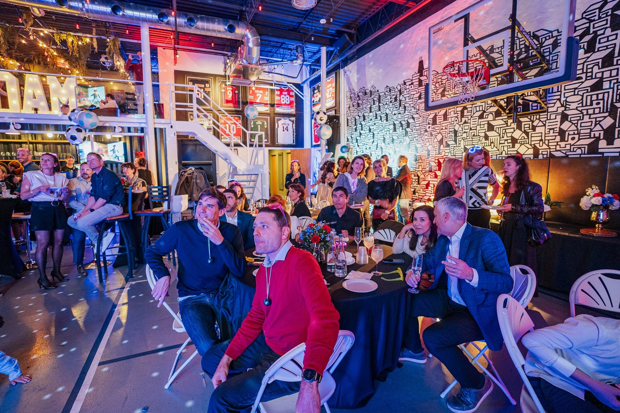 People are gathered in a lively indoor event space decorated with sports jerseys and football-themed decor, with some people sitting at tables and others standing and mingling.