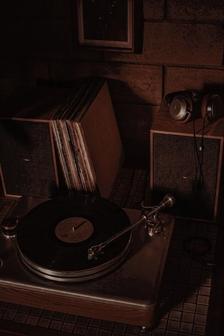 A vintage turntable playing a vinyl record, with speakers and a stack of records in a dimly lit room.