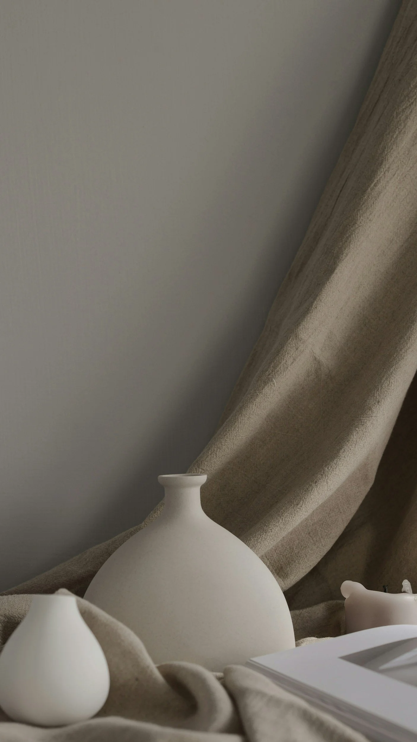 Minimalist display of ceramic vases with folded beige fabric and a book, against a plain wall.