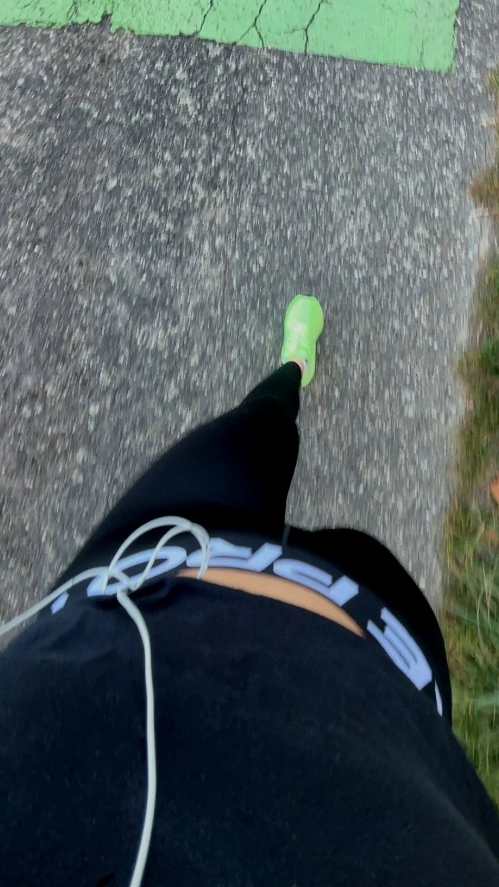 Photo prise du point de vue d'une personne en train de courir, portant des chaussures de course vertes, des vêtements noirs, et des écouteurs blancs, sur un trottoir en béton.