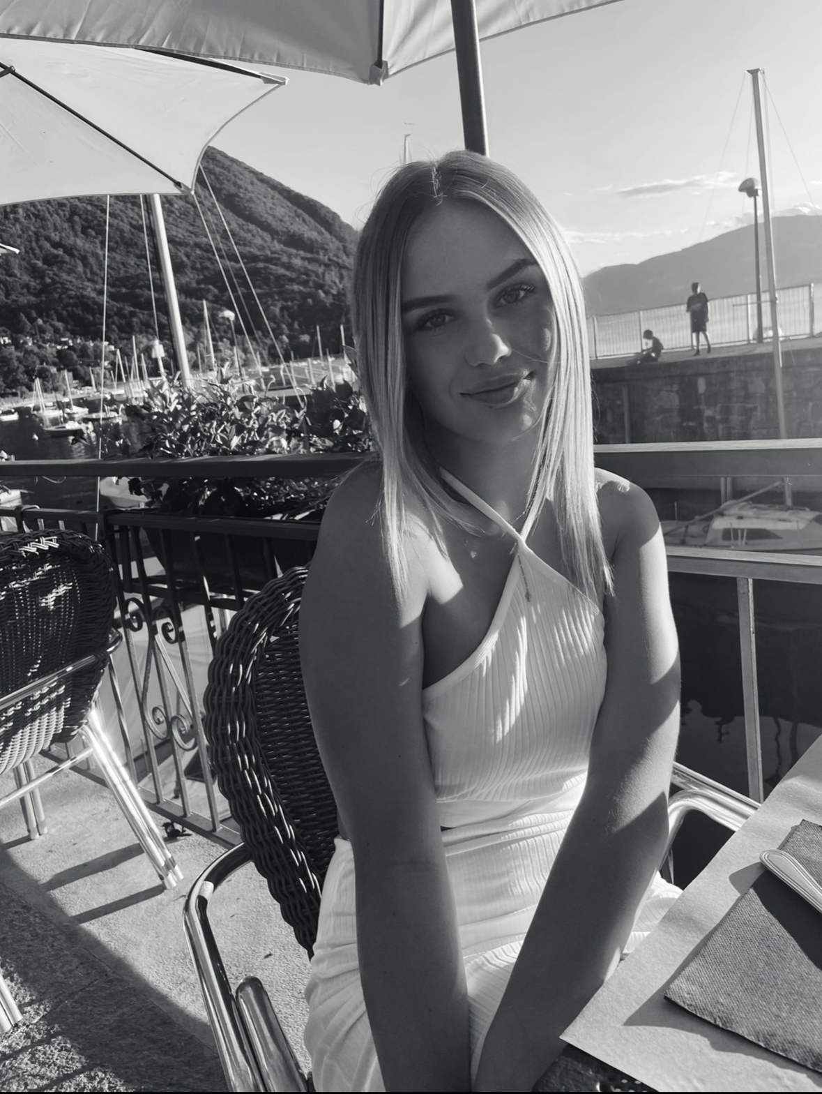 Jeune femme assise dans un restaurant en extérieur avec vue sur un lac et des montagnes, portant une robe à baillettes et un sourire, en noir et blanc.
