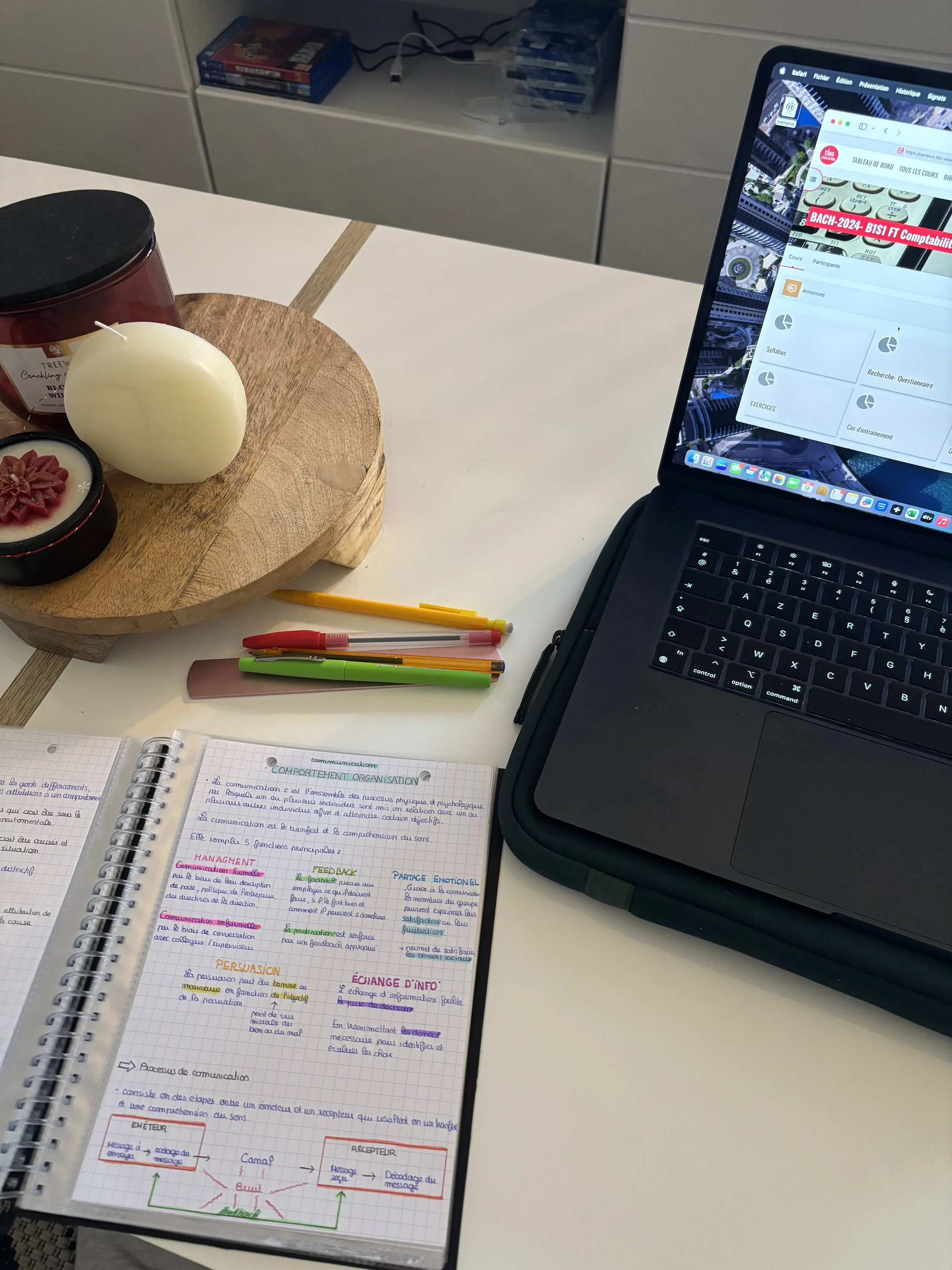 Page de notes écrites à la main sur un carnet, avec des titres en couleurs variées, à côté d'un ordinateur portable ouvert, sur une table blanche. Il y a aussi des stylos et des objets décoratifs comme une bougie blanche et une bougie parfumée.