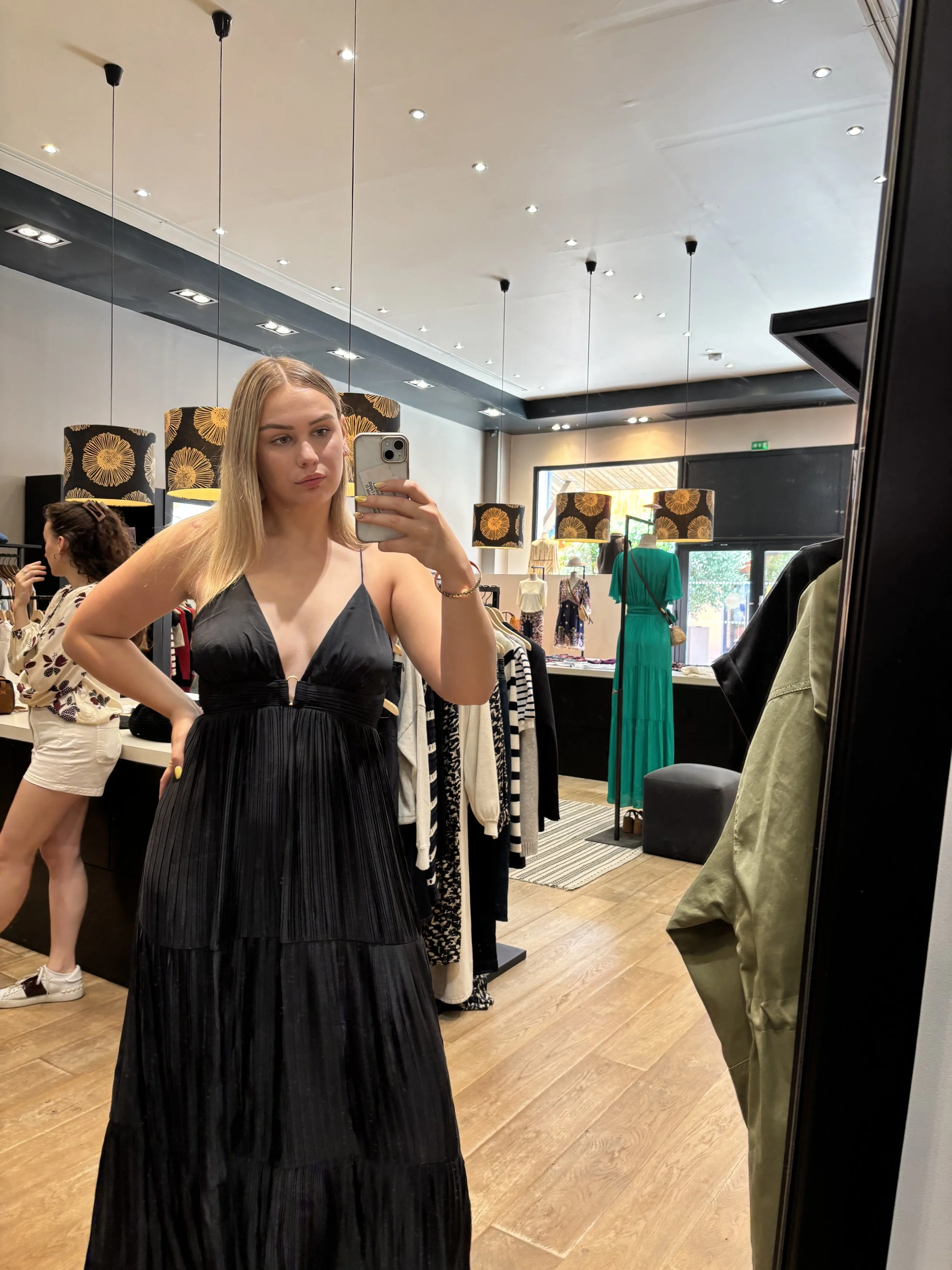 Une jeune femme blonde essaie une robe noire à rayures dans un magasin de vêtements, réfléchie dans un miroir.