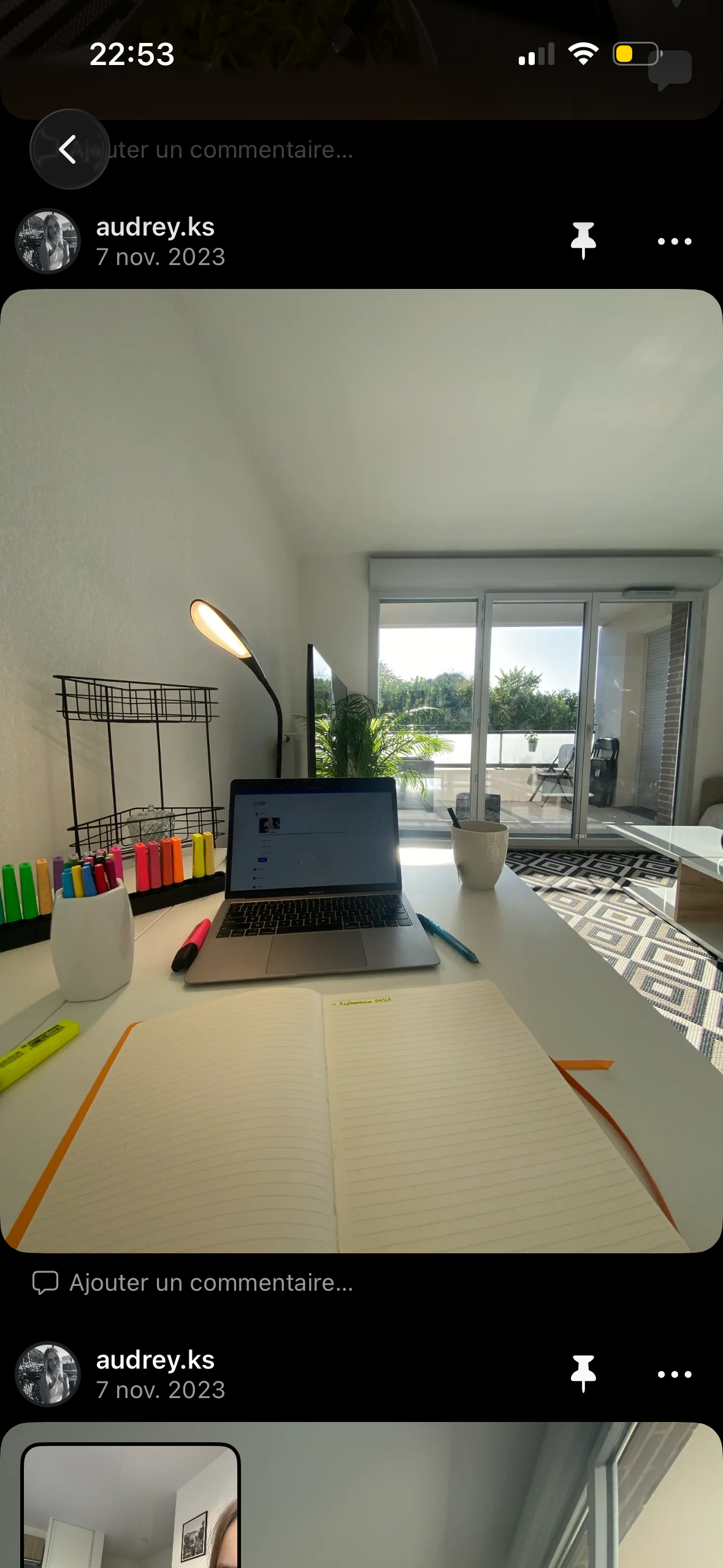 Bureau avec un ordinateur portable, un cahier ouvert, des marqueurs de différentes couleurs, une lampe de bureau, une tasse blanche, une plante verte. En arrière-plan, une grande baie vitrée donnant sur une terrasse extérieure avec du mobilier et la nature en arrière-plan.