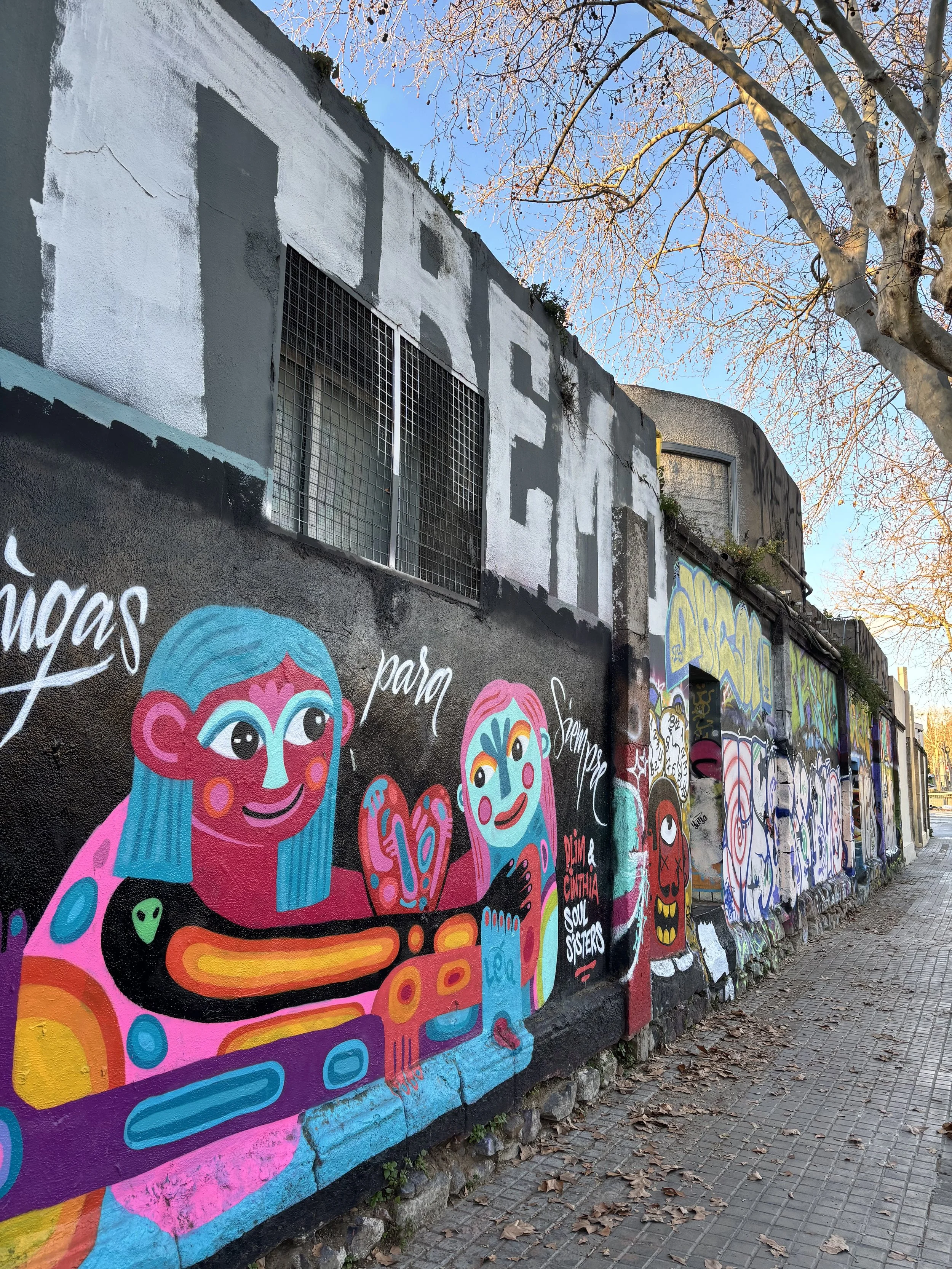 Mur de graffiti coloré avec illustrations de personnages stylisés et textes, avenue pavée avec feuilles mortes et arbres dénudés au-dessus, ciel clair.