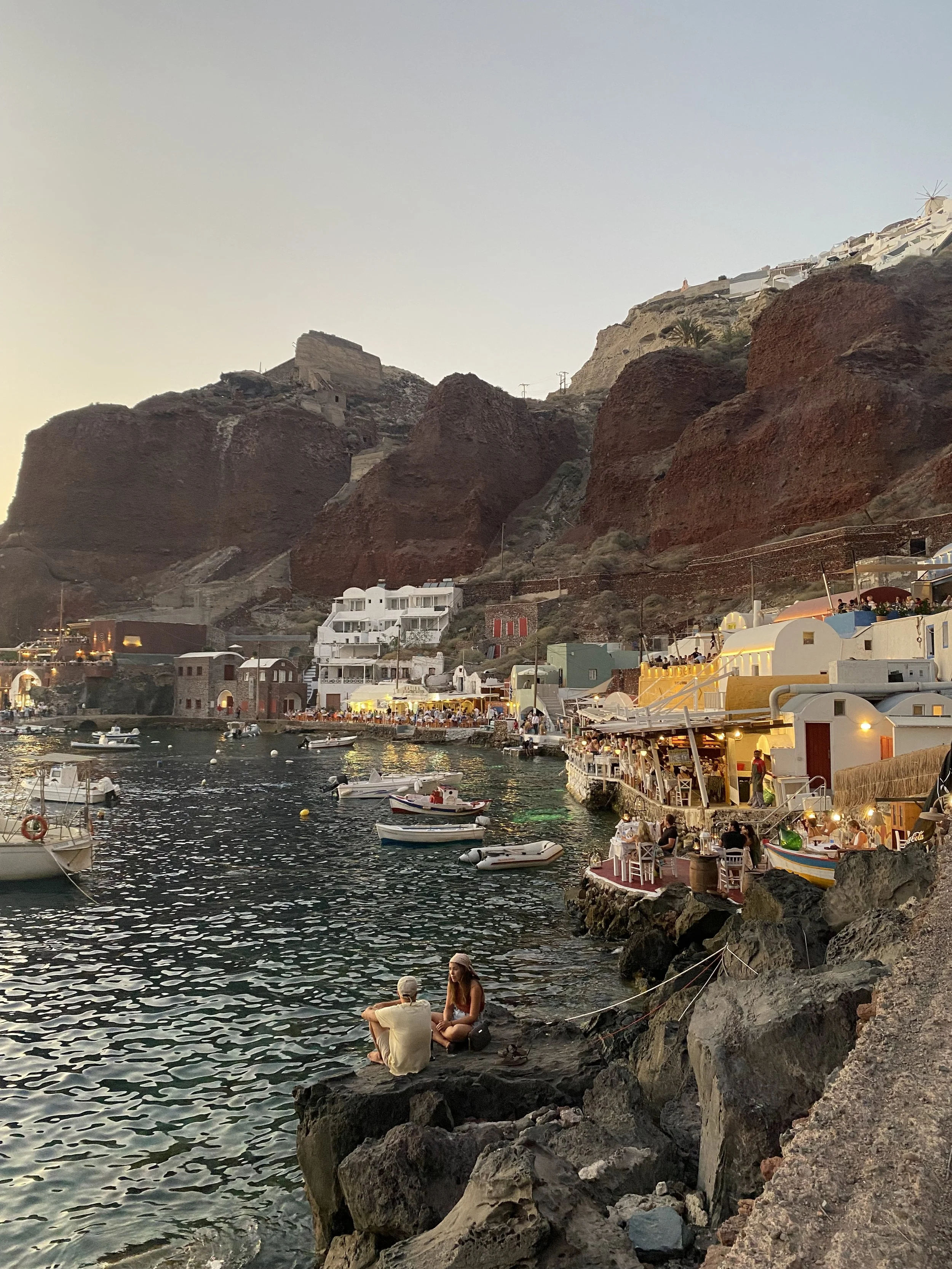 vue d'un port de pêche animé avec des bateaux, des bâtiments blancs et des restaurants en bord de mer, surplombés par une falaise escarpée avec des maisons blanches et une église, au coucher du soleil.