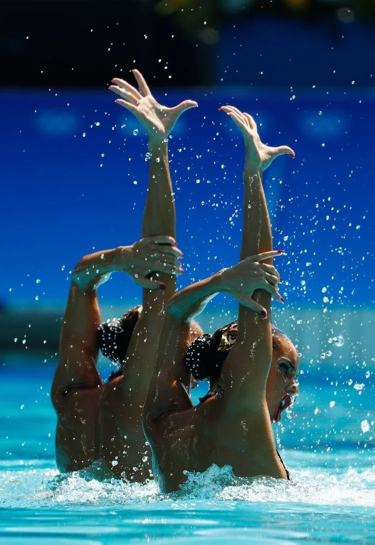Deux femmes pratiquant la natation synchronisée dans une piscine, effectuant une pose artistique avec leurs bras en l'air.