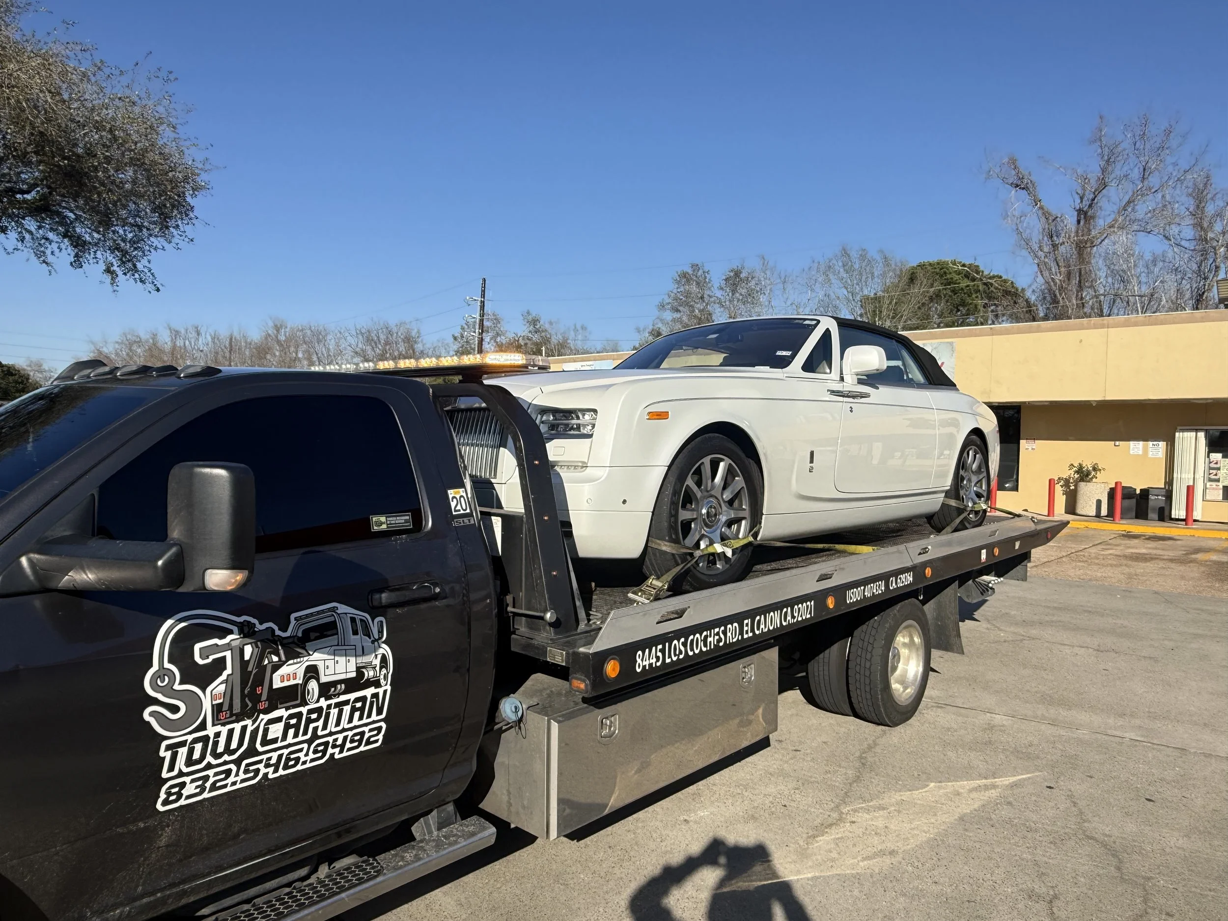 A white Rolls Royce  is being transported on a flatbed tow truck at night in front of a shopping plaza with stores