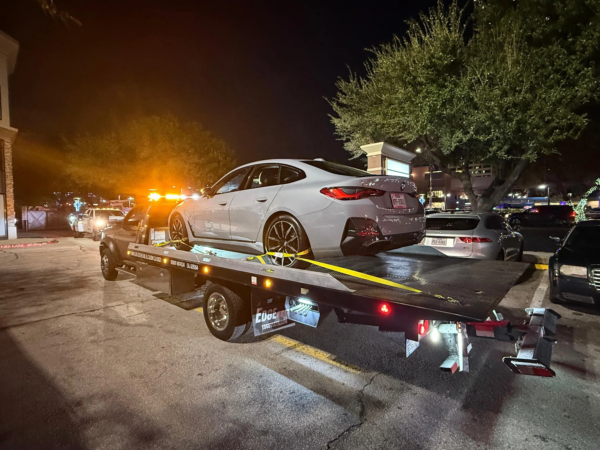 A silver luxury car loaded on a flatbed tow truck at night in a parking lot, with other parked cars and illuminated trees in the background.