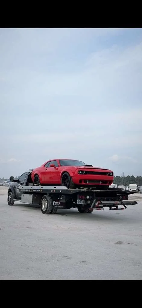 A red muscle car on a flatbed tow truck on a highway with vehicle traffic in the background under a cloudy sky.