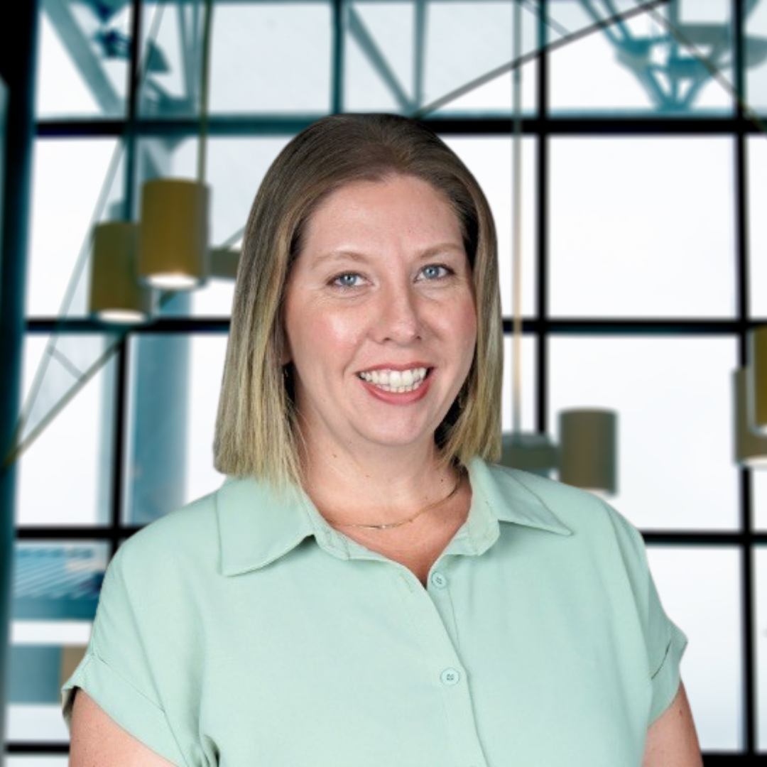Portrait of a smiling woman with shoulder-length hair, wearing a light green button-up shirt, standing in front of a large glass window with a modern building structure visible in the background.