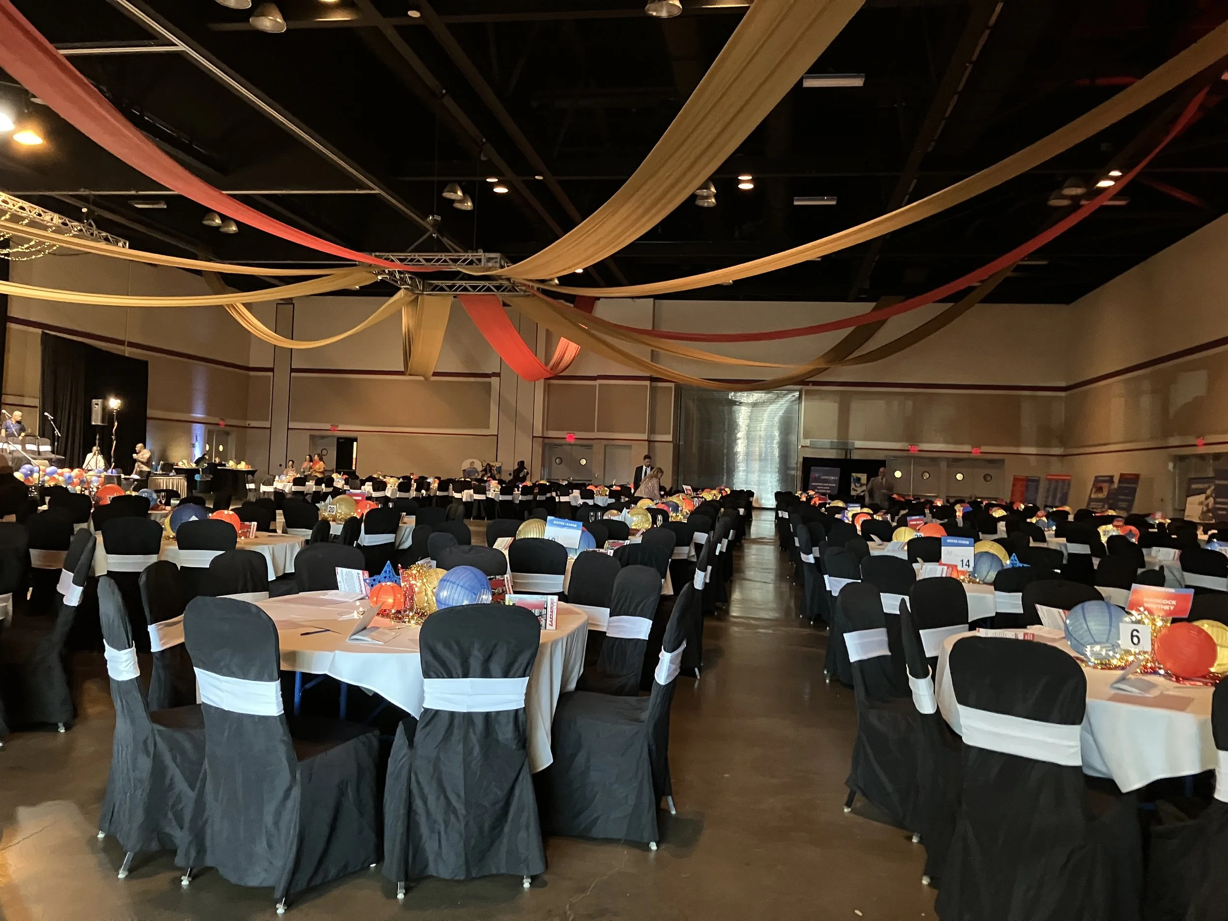 An event hall decorated with red, gold, and beige fabric drapes hanging from the ceiling, with round tables covered in white tablecloths and black chair covers with white sashes. The tables are decorated with colorful paper lanterns, illuminated deco