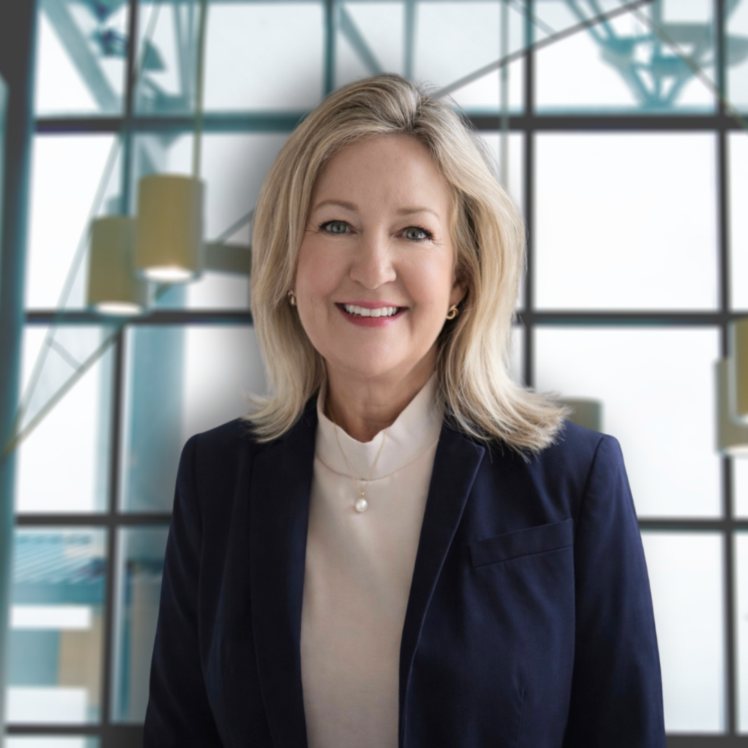 A smiling blonde woman in a navy blazer and white blouse stands in front of a modern glass building interior.