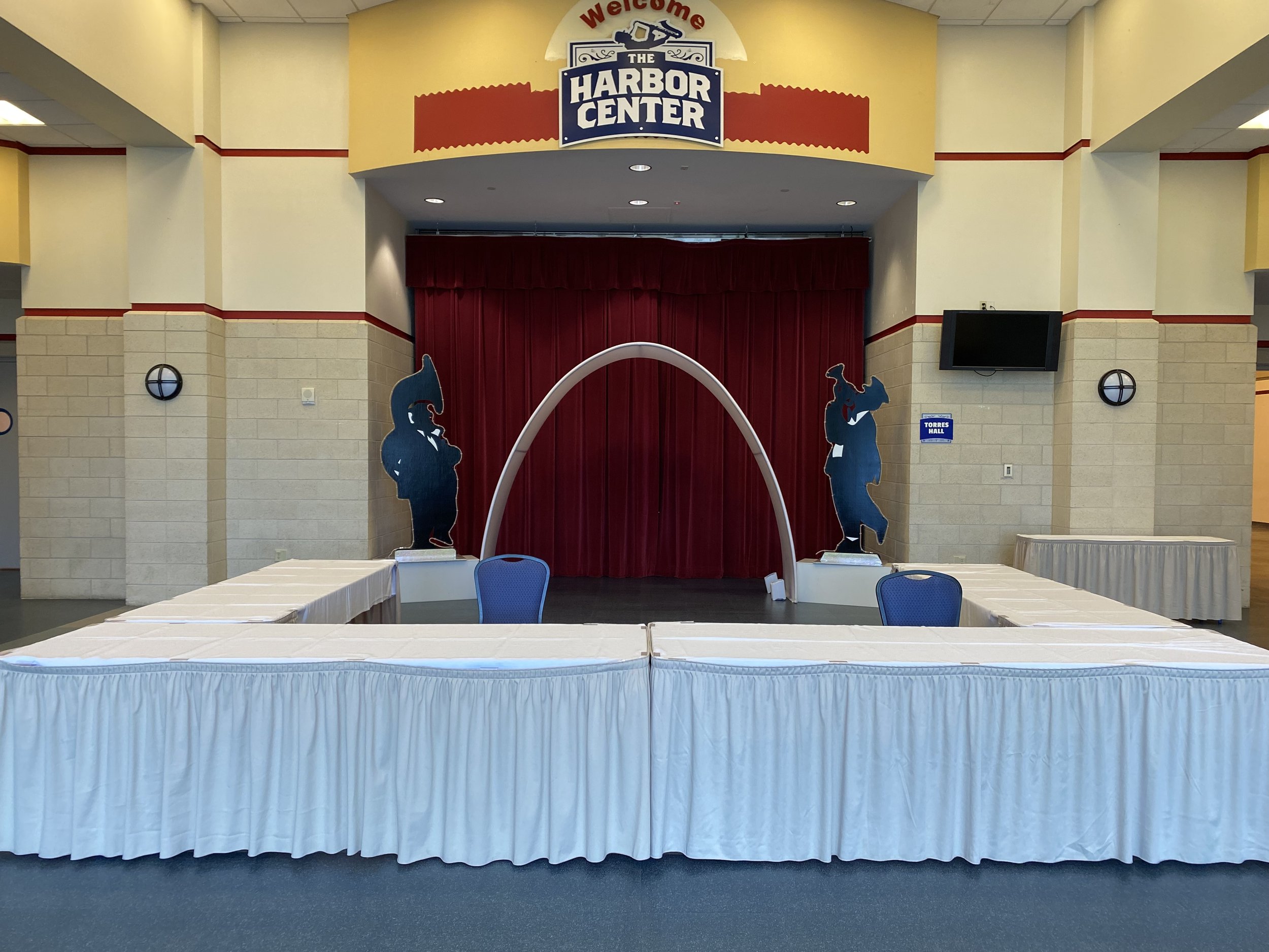 Empty event stage at Harbor Center with red curtains, two pirate-themed silhouettes, a white arch, and a white skirted table in front.