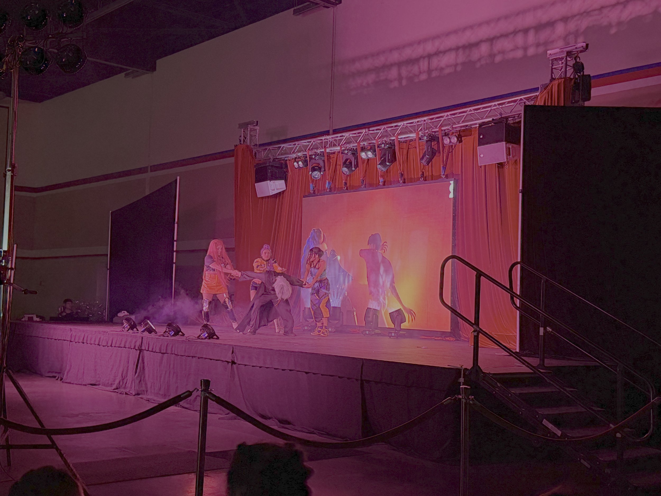 Stage with performers, large screen, stage lighting, and curtains, in a dimly lit auditorium.
