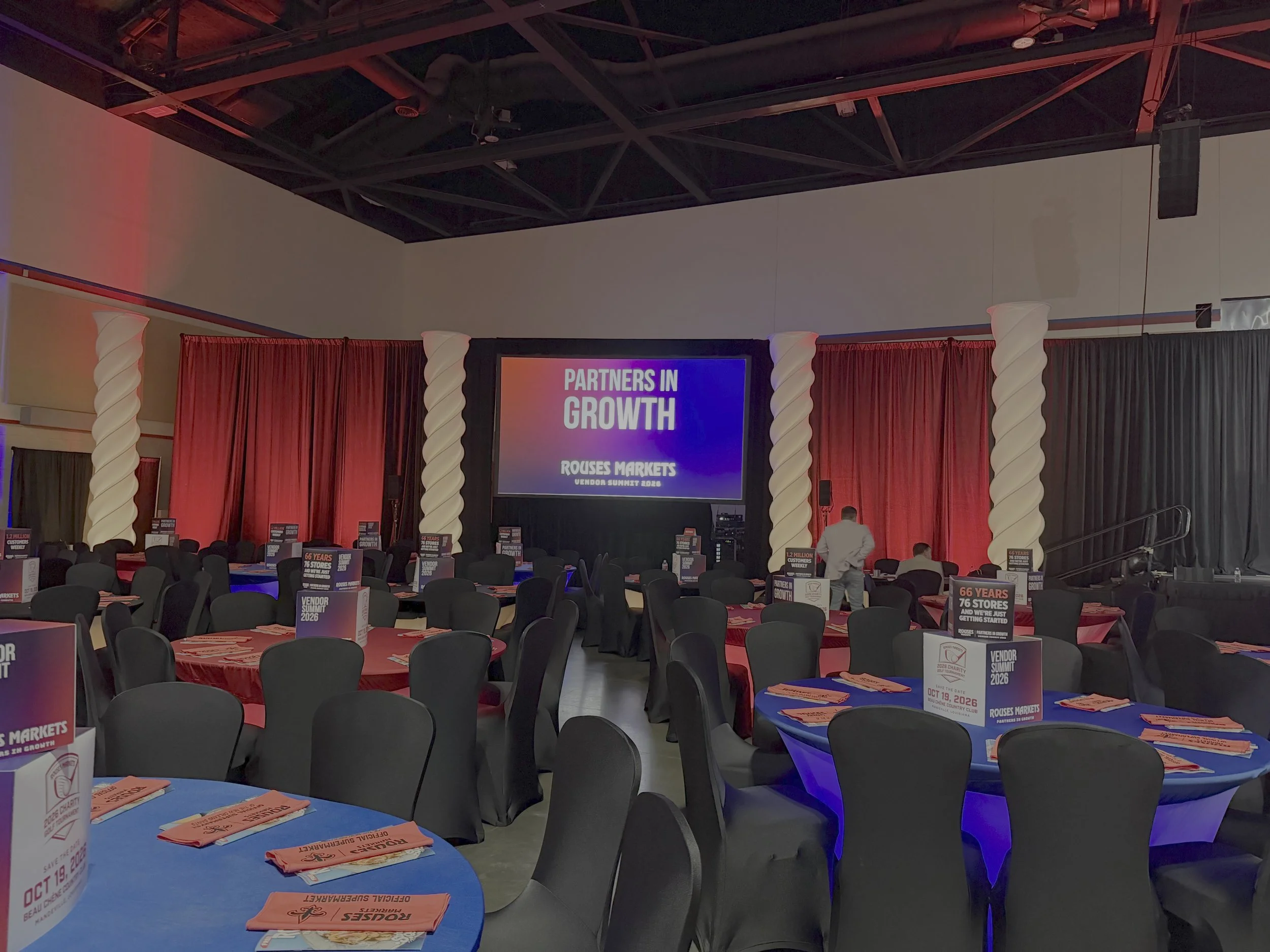 Conference room set up with round tables covered with blue tablecloths. Each table has stacks of orange pamphlets and a white cup. Several black chairs surround the tables. A large screen in the background displays the text "Partners in Growth" and "