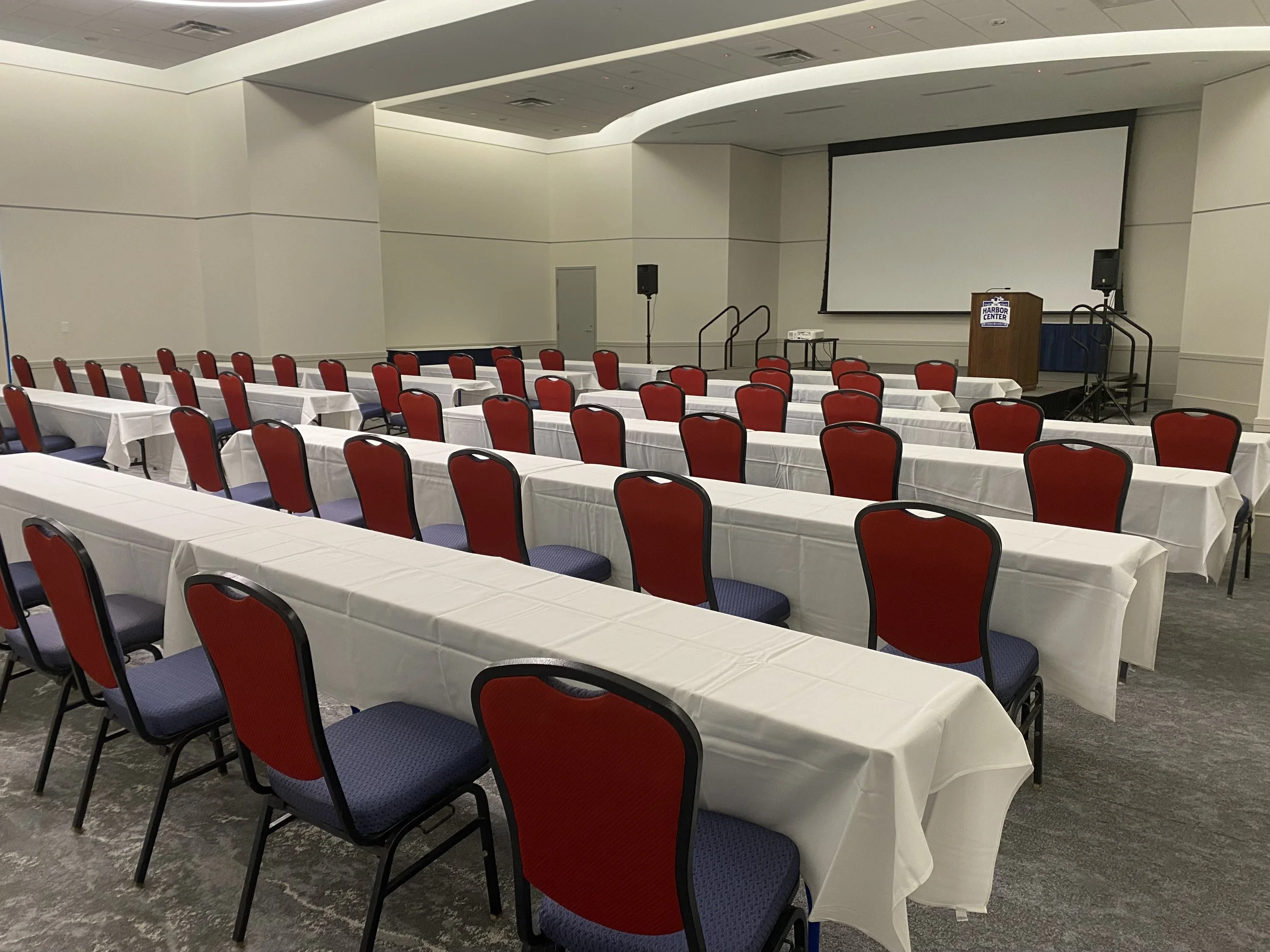 Conference room set up with rows of tables covered in white tablecloths and red chairs, facing a stage with a projector screen and a podium.