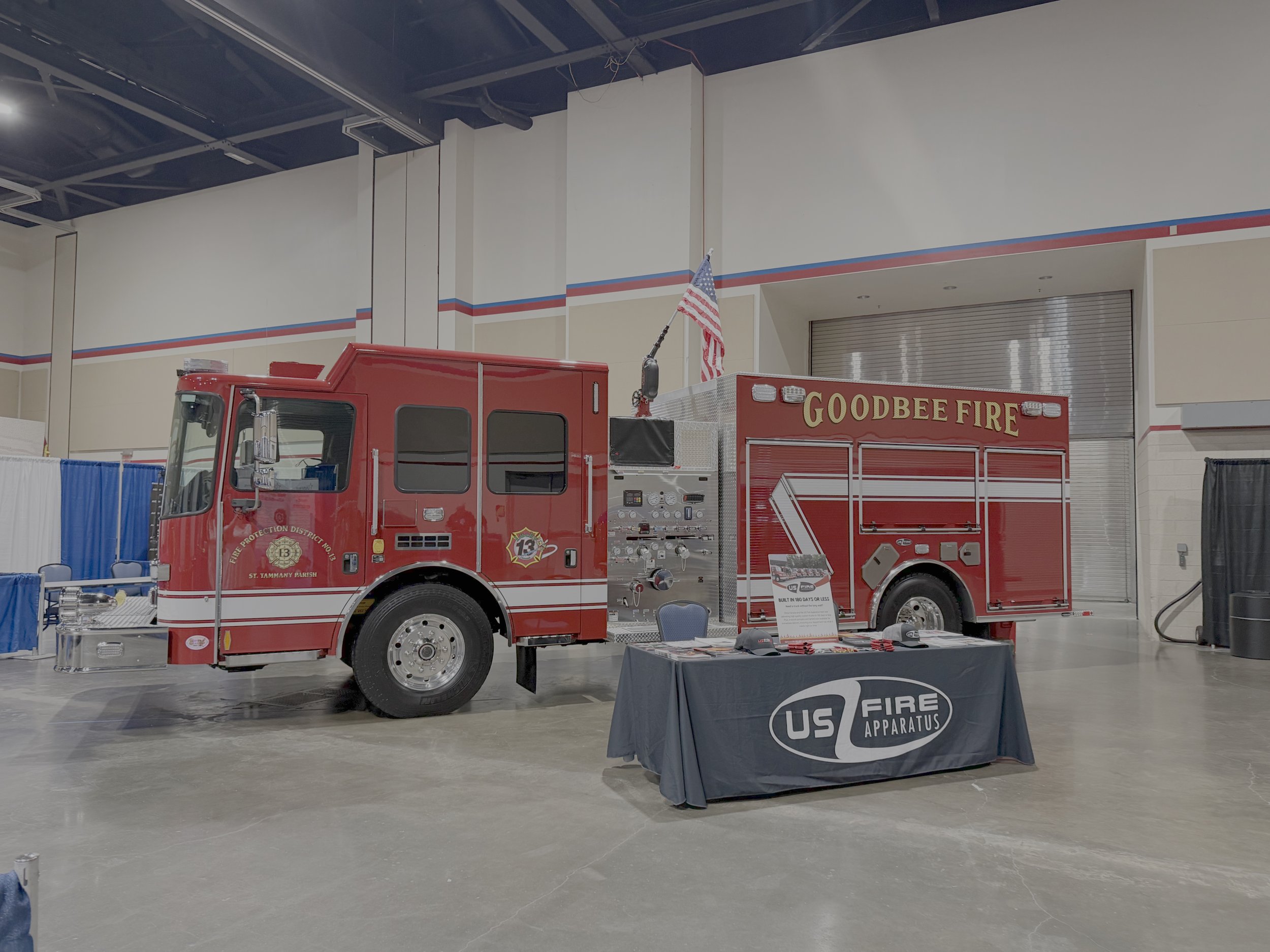 Red fire truck labeled 'Goodbee Fire' on display at an indoor event with a table covered in a black tablecloth that reads 'US Fire Apparatus' and an American flag on top of the truck. Blue and white curtains with a high ceiling and industrial lightin