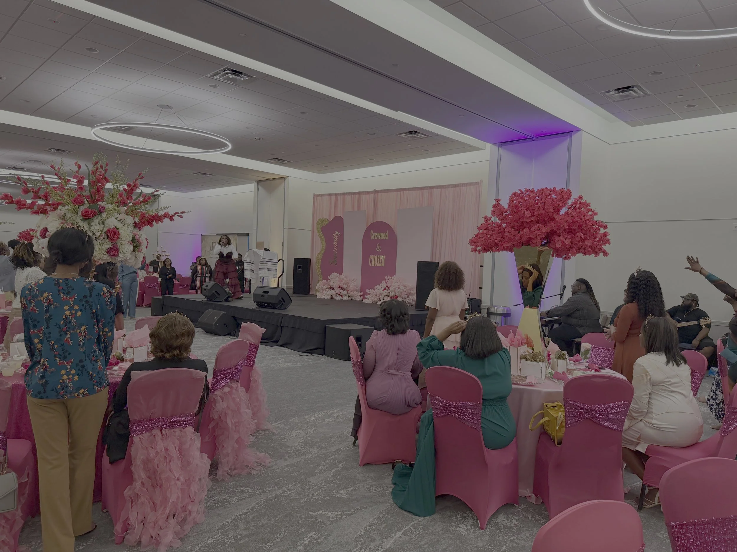 A large indoor event space decorated with pink accents, including pink chair covers, pink floral arrangements, and pink balloons, with various people seated and standing around, some on a stage at the front.