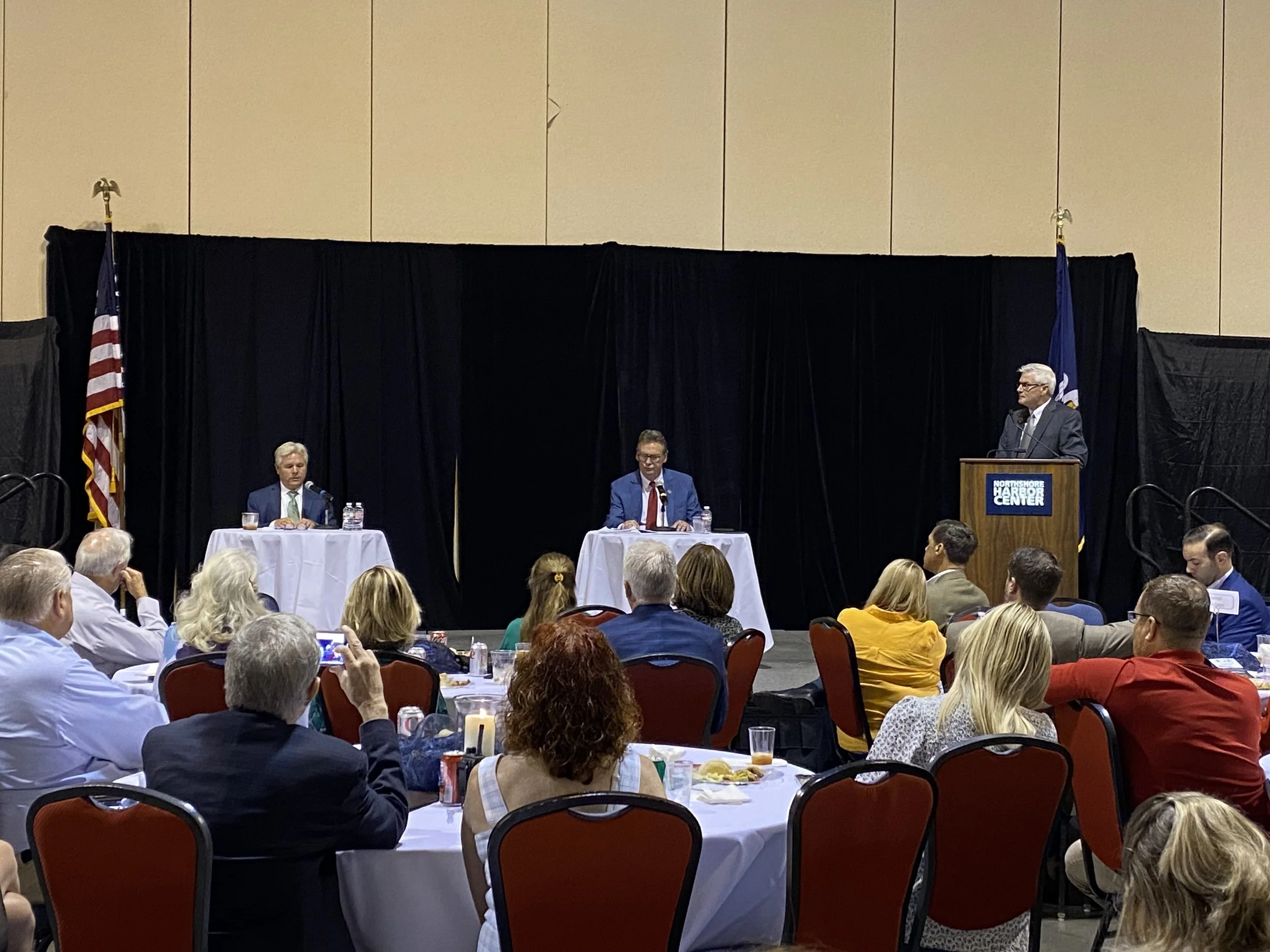 A panel discussion at the Northshore Harbor Center with three men, two seated at tables on the stage and one standing at a podium, with an audience watching.