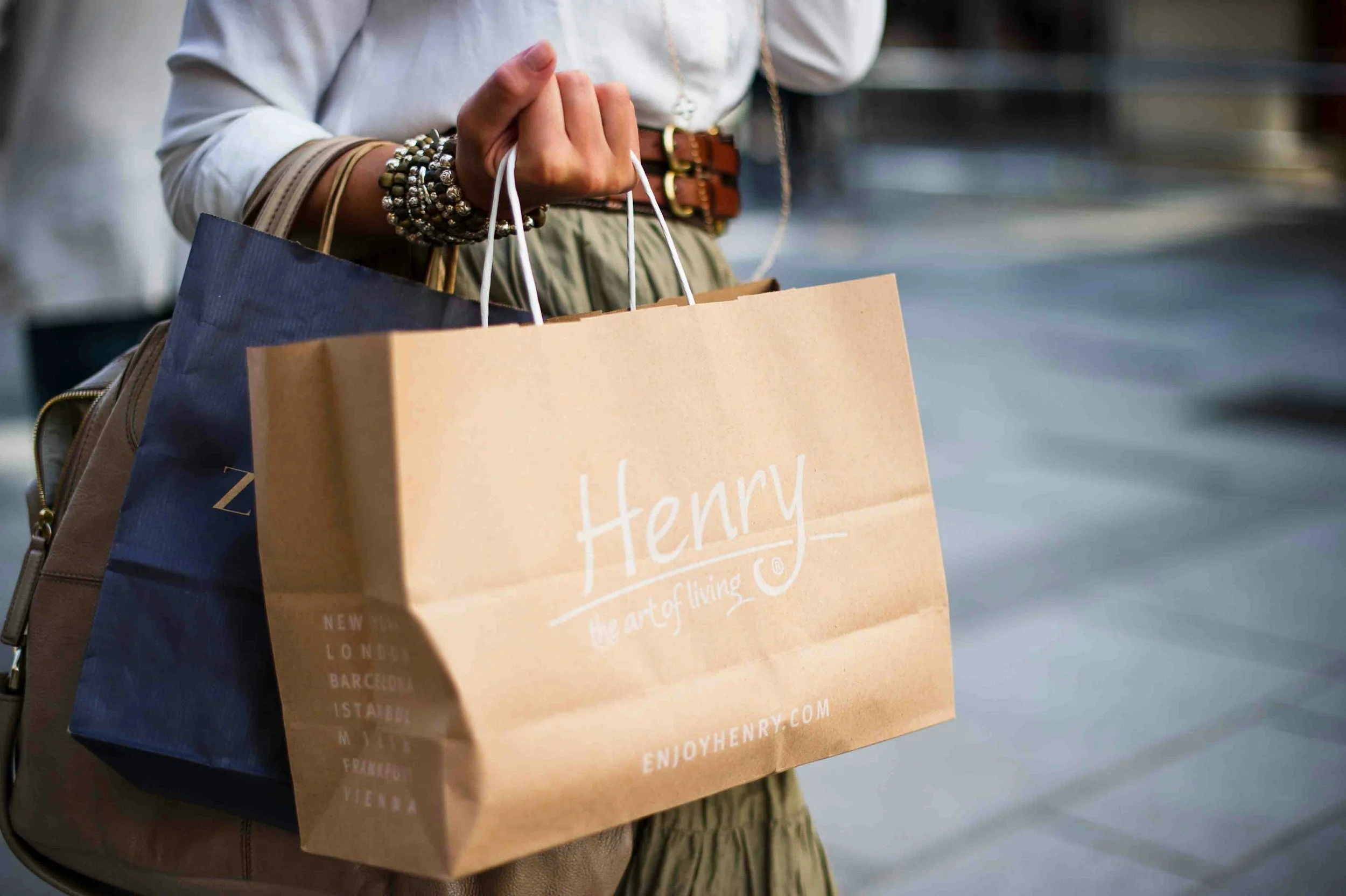 Person holding shopping bags, including a tan one with 'Henry' and other bags behind it, standing on a city sidewalk.