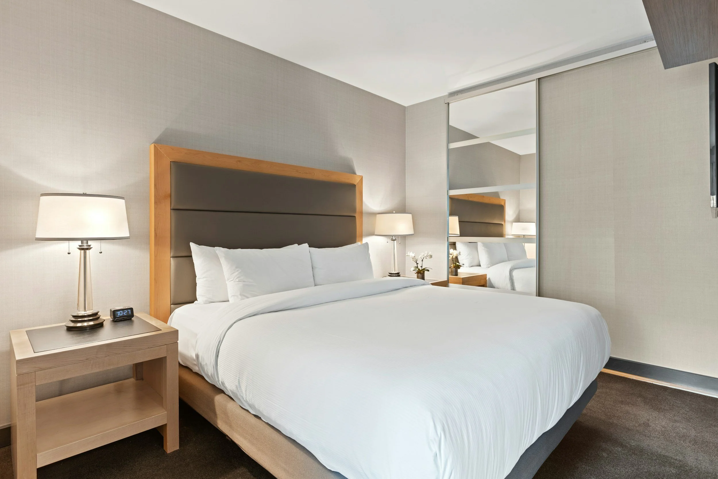 A modern hotel bedroom with a large bed, nightstands, lamps, and mirrored closet doors.